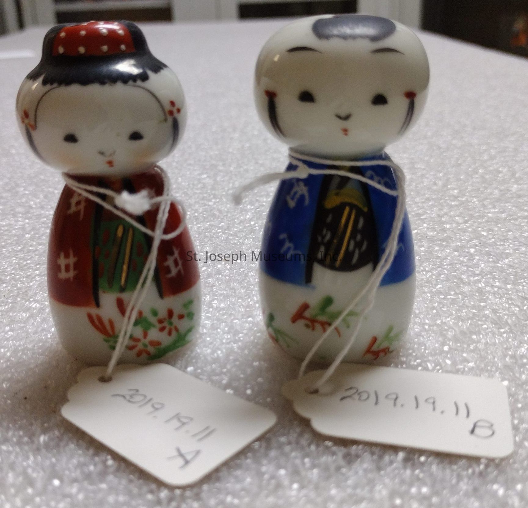 The image displays two small, intricately painted kokeshi dolls placed side by side on a textured surface, likely made of polystyrene. Each doll exhibits a unique, hand-painted design; the doll on the left is adorned in a red and green ensemble, while the doll on the right sports a blue and white attire, both featuring delicate floral and traditional patterns. They have simple, charming facial expressions characterized by small eyes and gentle smiles. Both dolls have a string attached around their necks, leading to tags inscribed with “2019.19.11” followed by “A” and “B” respectively, suggesting they might be part of a collection or exhibit. The background seems neutral, possibly suggesting a display or an exhibition setting, effectively emphasizing the cultural and artistic details of these dolls.