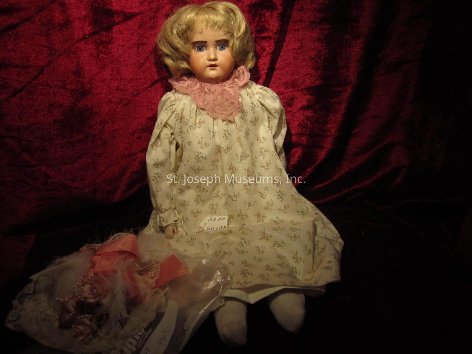 The image depicts a vintage doll with blonde hair and striking blue eyes, dressed in a floral-patterned gown with a pink lace collar. The doll is positioned against a rich red velvet background, which enhances its antique and somewhat mysterious aura. The fabric of the dress appears to be delicate, showcasing small floral prints, indicative of a bygone era. To the side of the doll, there is a small plastic bag containing additional items, possibly accessories or related objects. There is a price tag visible on the doll, suggesting it might be part of a collection or on display for sale. The overall ambiance of the image is nostalgic, capturing the essence of historical craftsmanship in doll-making. The lighting is dim, casting soft shadows that further add to the timeless and somewhat eerie atmosphere surrounding the doll, drawing attention to its intricately painted facial features and the detailed artistry of its attire, making it a captivating subject for collectors and enthusiasts of vintage items.