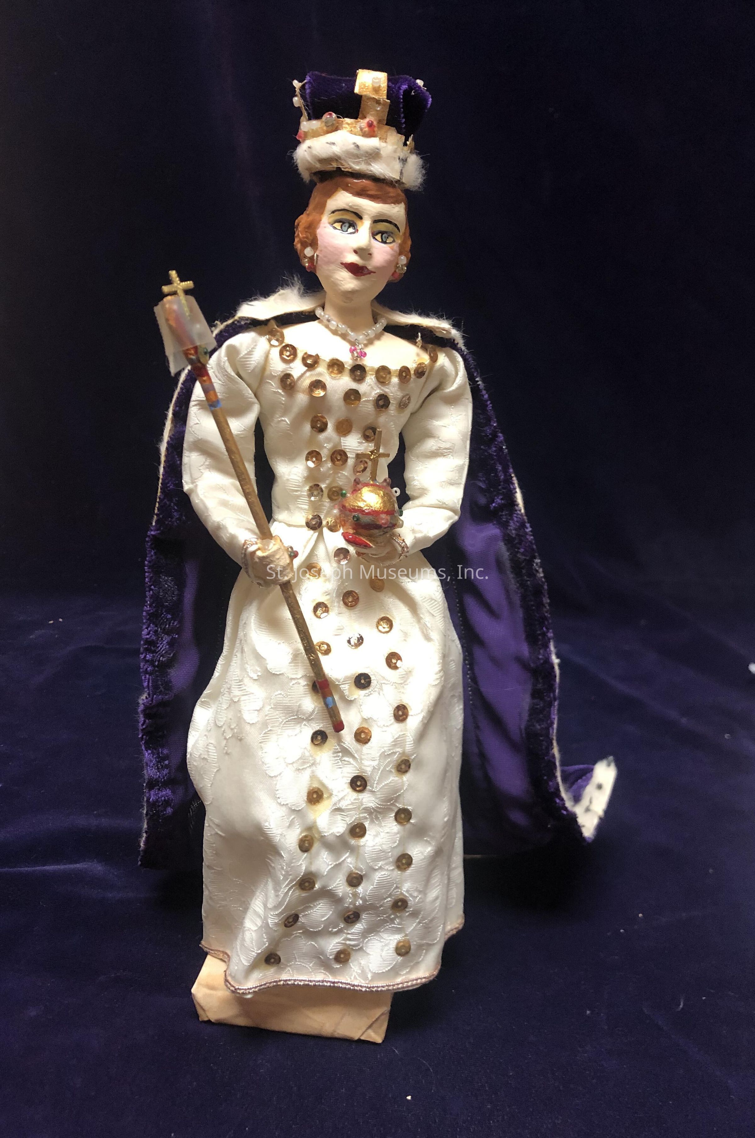 The image shows a handcrafted doll adorned in regal attire, featuring a detailed white gown embellished with gold sequins and floral patterns. The doll wears a lavish crown on its head, decorated with jewels and a plush trim, while holding a golden orb and a scepter in its hands. A rich purple velvet cape with an ermine-like white lining drapes elegantly from the shoulders, enhancing the doll's royal appearance. The facial features are meticulously painted, with bold eyebrows, expressive eyes, and red lips. The background is a deep shade of blue, providing a striking contrast that draws attention to the ornate and intricately designed costume of the doll.