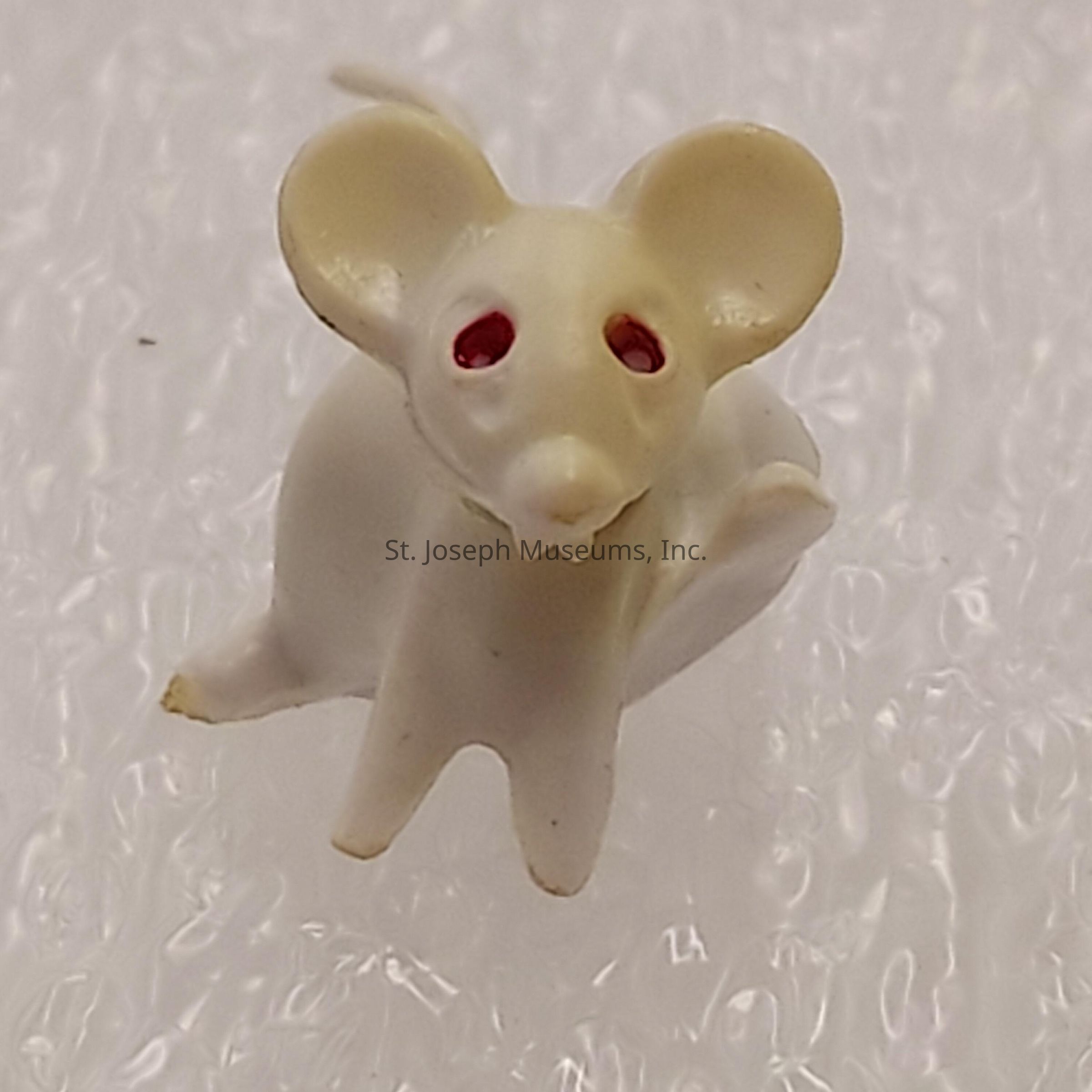 The image shows a small, white figurine resembling a mouse with large, rounded ears, pointed snout, and two red eyes, positioned on a textured, slightly reflective surface that appears to be bubble wrap. The mouse's head is slightly tilted, giving it an inquisitive expression. The light source emphasizes its glossy texture, casting soft shadows around the figurine's contours, particularly underneath its ears and along its body. The red eyes stand out against the white body, adding a striking contrast, while the sculpture captures a simplified and stylized representation of a mouse, focusing more on essential features for identification rather than on intricate details. This simplicity contributes to its charming and whimsical appearance, appealing to viewers with its cute and endearing design. The smooth finish of the figurine complements the intricate pattern of the background, creating an engaging visual contrast between the subject and its environment. The overall composition is minimalistic yet effectively draws attention to the central figure.