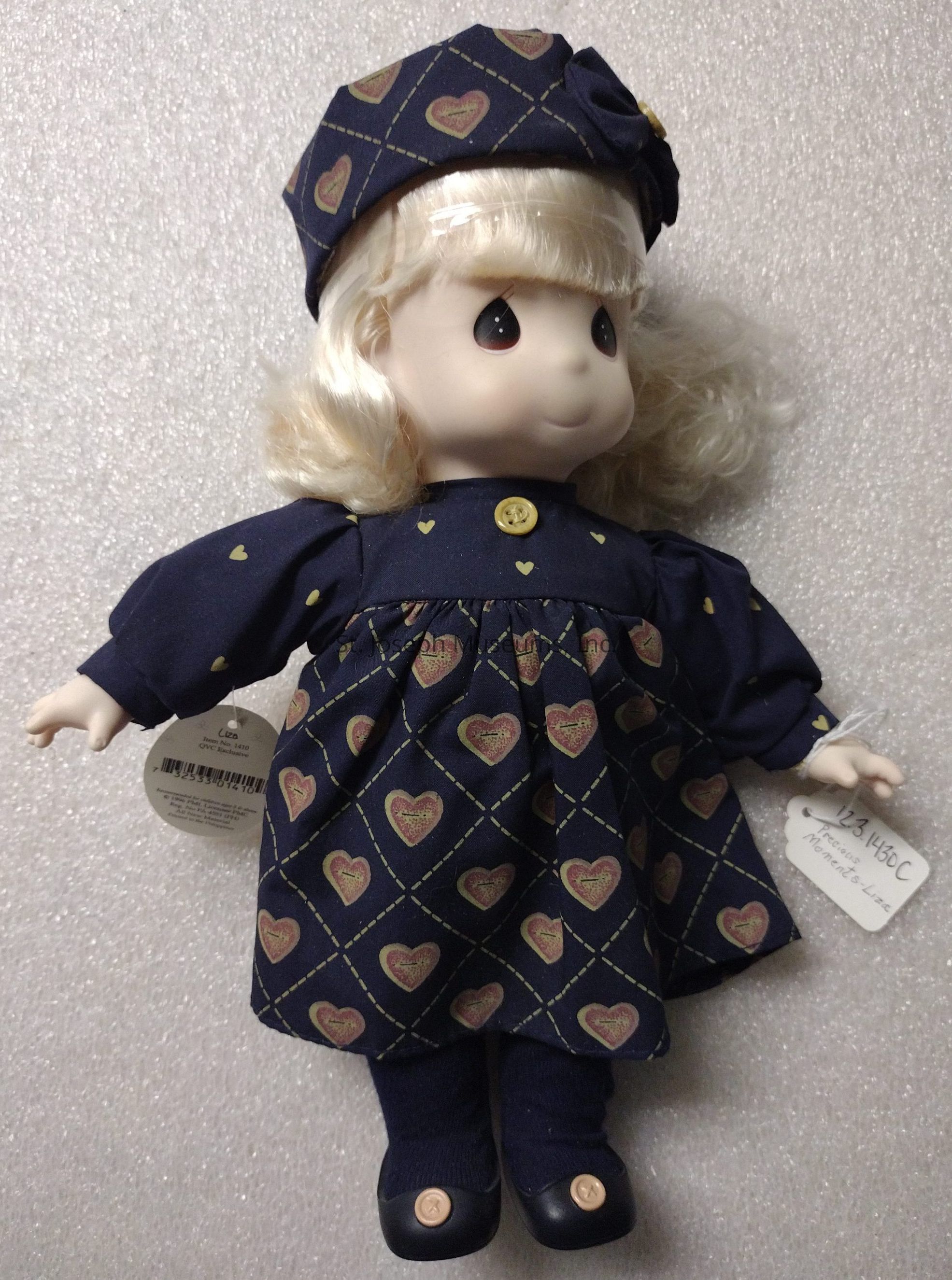 The image displays a charming doll with a porcelain-like complexion, adorned with soft, curly blonde hair that gently frames its rounded face. The doll is dressed in a navy blue ensemble featuring a dress embellished with a pattern of hearts set against a subtle diamond grid, and accessorized with a matching hat. A solitary yellow button securely fastens the dress at the collar, complementing the small yellow hearts scattered across the fabric. The doll wears navy stockings and black shoes with decorative buttons, completing its outfit. Hanging from its outfit are two tags, positioned on either side of the body, hinting at its authenticity or collectible nature. The eyes are large and expressive, giving the doll a sweet and innocent expression that adds to its overall endearing and collectible appeal.