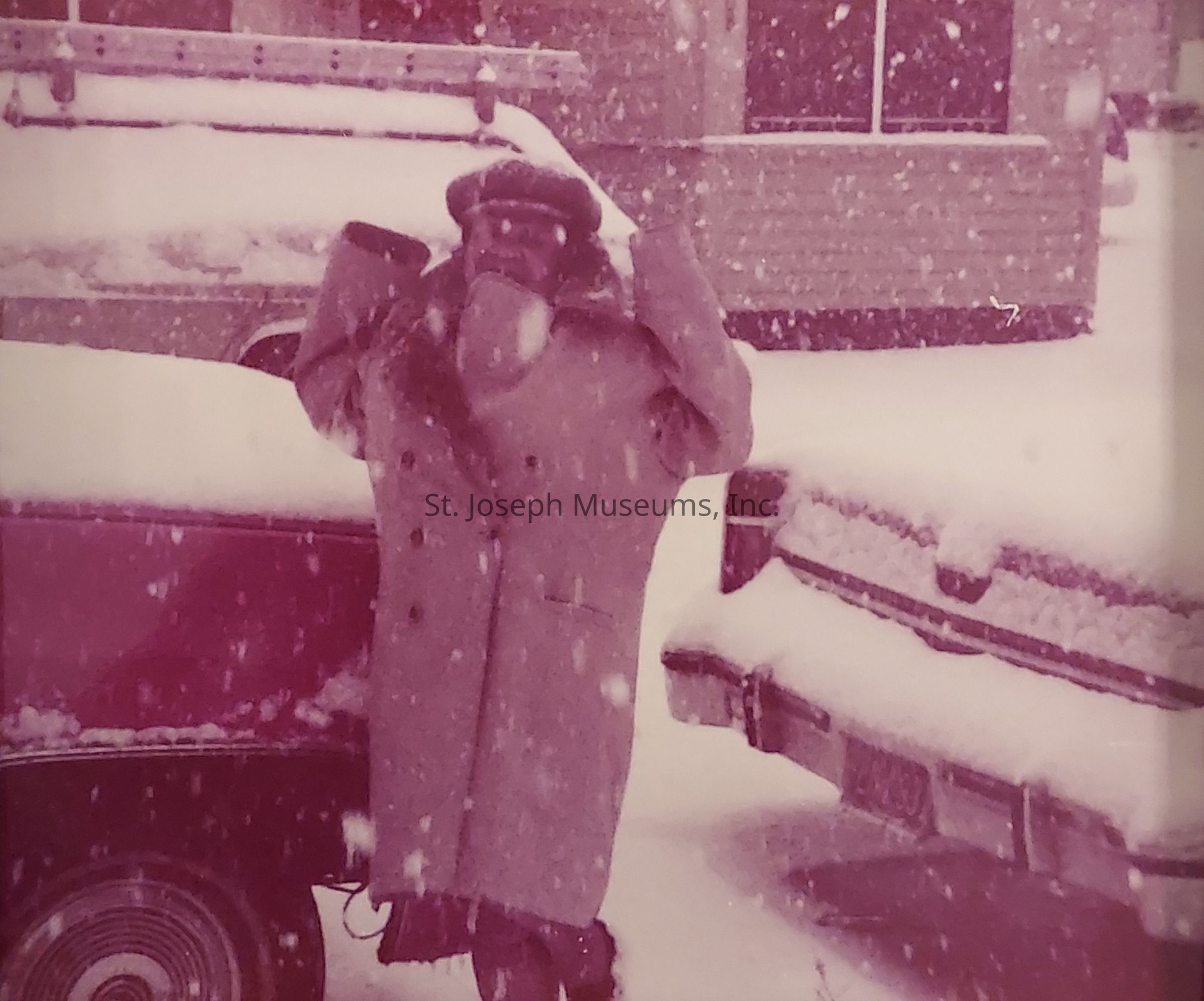 The image captures a person standing amidst a heavy snowfall, dressed in a long, thick, double-breasted coat, with a cap pulled low on their head and a scarf wrapped around their face for protection against the cold. Snow blankets the surrounding cars, forming a thick layer on their roofs and hoods, while the vehicle's tires are partially obscured by the accumulating snow. A ladder is visibly secured on top of one of the cars, hinting at possible outdoor work despite the weather conditions. The scene has a vintage feel, with a pinkish tint affecting the overall color of the photograph, likely due to fading over time. The heavily falling snowflakes add a dynamic element to the composition, emphasizing the wintry conditions and the person's preparedness for the harsh environment. The background shows a building with horizontal siding, partially obscured by the storm, further highlighting the intensity of the snowfall. This evocative image captures a moment of perseverance and adaptation in the face of nature’s challenges.