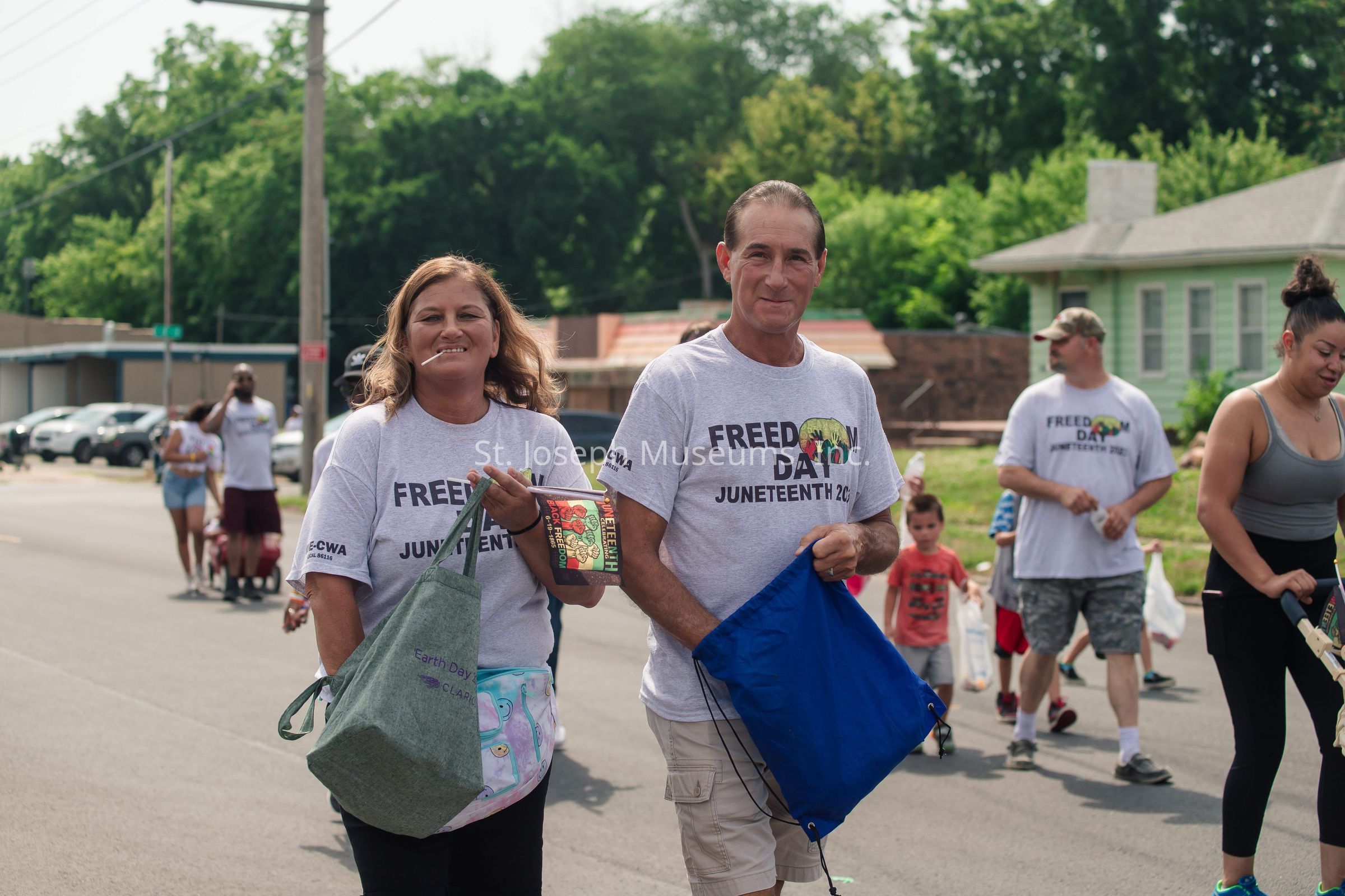 In the image, a group of people is participating in a street event, wearing matching shirts with the words "Freedom Day Juneteenth" printed on them, indicating a celebration of Juneteenth. A woman on the left is smiling with a lollipop in her mouth and holding a reusable bag, while the man next to her is carrying a blue drawstring bag and smiling at the camera. The background reveals additional participants, including a man and woman on the right and several children, some of whom appear to be carrying bags, suggesting activities or a parade. The setting is a suburban area with cars parked along the street and houses surrounded by greenery. The atmosphere seems festive and communal, with people casually walking and engaging in what appears to be a joyous and meaningful commemorative event.