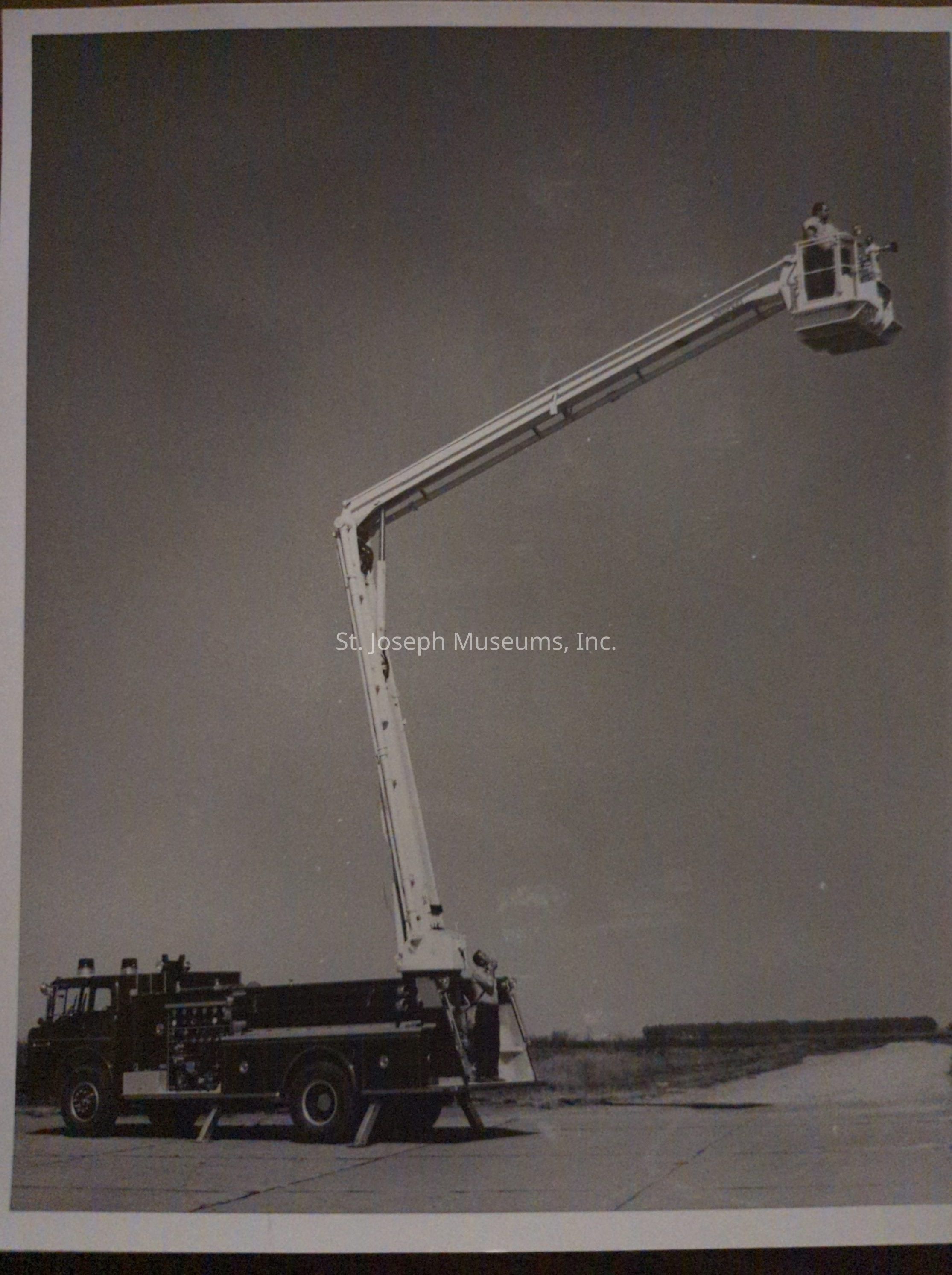 Black-and-white photograph depicting a fire truck from Snorkel Co., positioned on Lake Road and Arizona, featuring an extended aerial platform. A person operates the platform high above, showcasing its impressive reach and functionality, with the truck placed on a flat road, illustrating its readiness for firefighting operations.