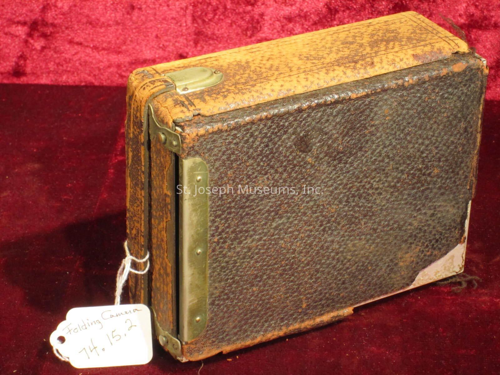 The image shows a vintage folding camera placed against a rich, burgundy background. The camera features a worn and textured leather exterior with visible signs of age and use, giving it a rustic and historical appearance. Metal components can be seen along the edges, suggesting durability and functionality. A small white tag is attached to the camera with a string, inscribed with the words "Folding Camera 74. 15. 2," indicating its categorization or inventory number. The overall composition highlights the camera's antique charm and craftsmanship, evoking a sense of nostalgia and appreciation for early photographic technology.