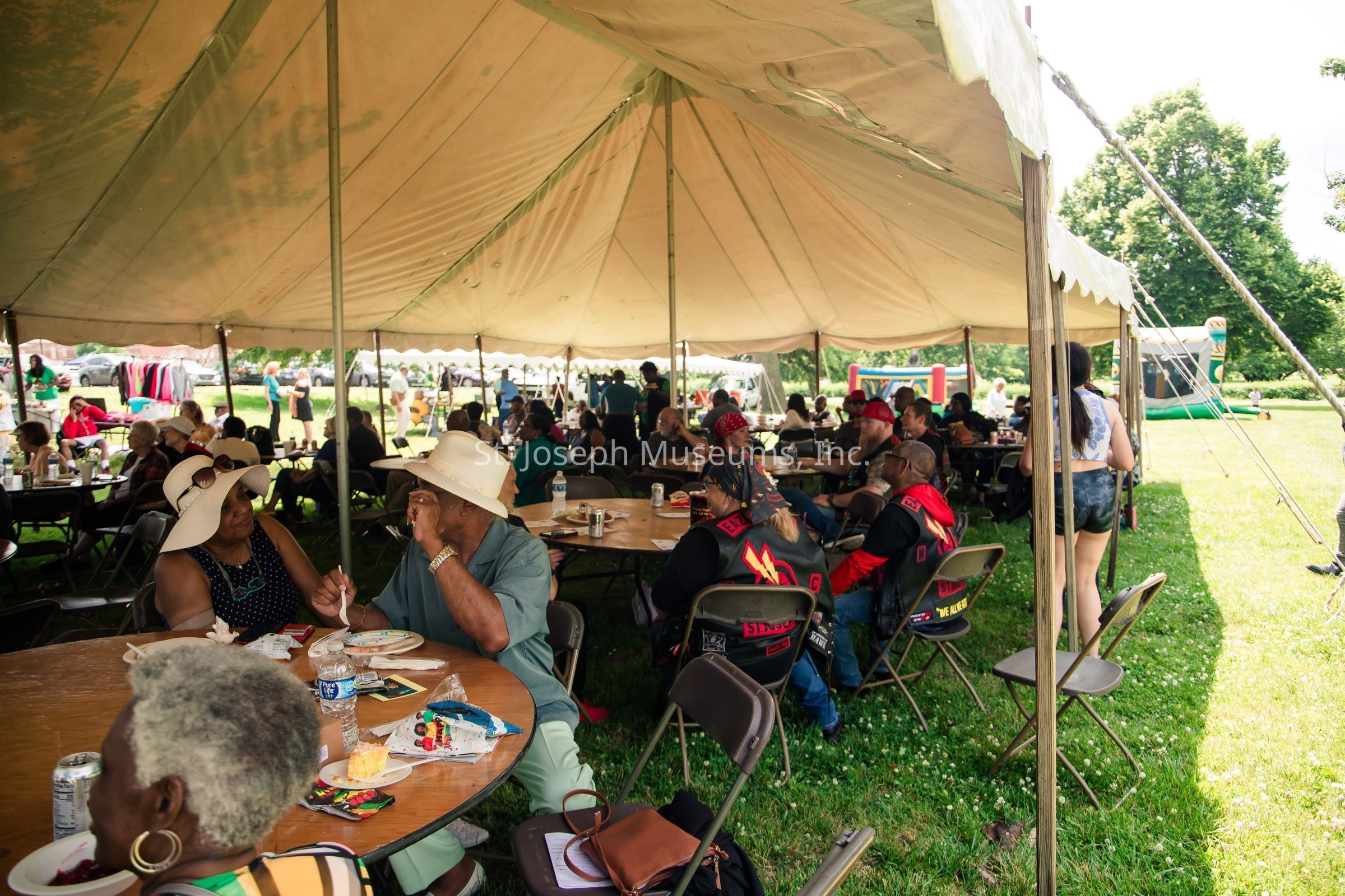 The image depicts a lively outdoor gathering under a large canopy tent, where numerous people are seated at round tables, engaging in conversation and enjoying food. Attendees of varying ages appear relaxed and content, suggesting a community event or celebration. The tables are adorned with plates, cups, and snacks, while some individuals wear wide-brimmed hats, possibly to shield themselves from the bright daylight. In the background, more people are visible outside the tent, along with inflatable play structures hinting at family-friendly activities. The grassy setting and surrounding trees contribute to the scene's warm and inviting atmosphere.