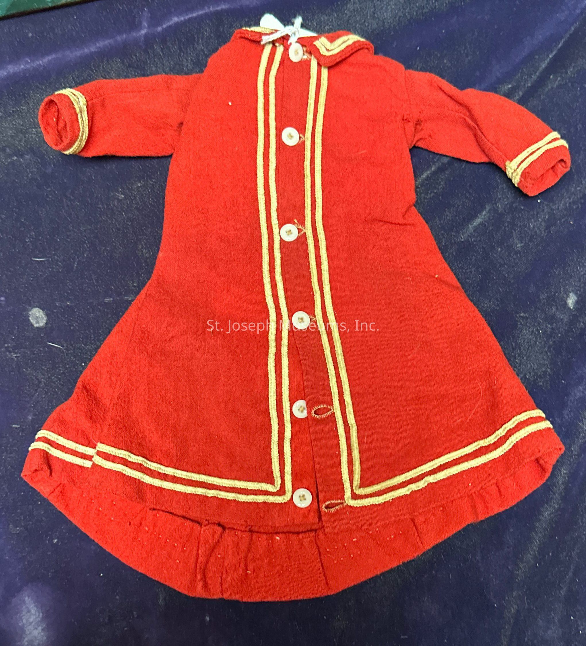 The image shows a vintage red coat made from a thick, textured fabric laid out on a dark purple surface. The coat features a series of white buttons running centrally from the collar to the hem. It has long sleeves and gold trim detailing along the button path, at the cuffs, and around the hem, adding a touch of elegance and contrast to the deep red material. The outfit appears to be designed for a small child or a doll, given its size and the style of the garment, which evokes a nostalgic charm. The setting provides a simple yet rich backdrop, accentuating the vibrant colors and intricate details of the piece.