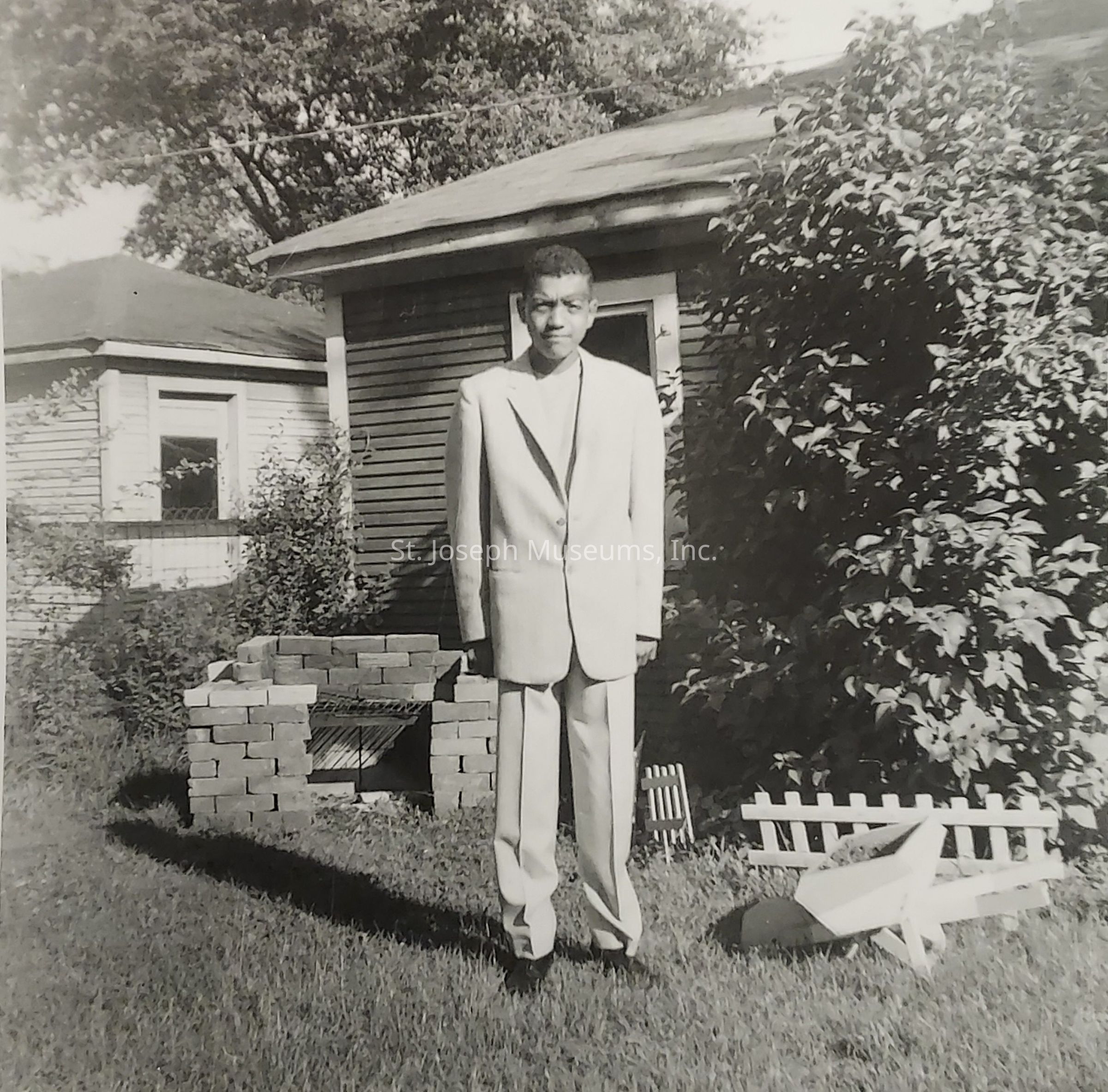 In this black and white photograph, a young man stands in a grassy backyard area, dressed in a light-colored suit. He appears poised and confident, positioned centrally in the frame. Behind him, a small building, likely a shed or garage, is partially obscured by bushes and vegetation, adding an element of depth to the composition. To his left, a simple brick structure, perhaps a rudimentary barbecue grill, lines up neatly, while to his right, a small white wooden picket fence and model airplane add a touch of nostalgia and innocence to the scene. The trees and shrubbery surrounding the area create a sense of enclosure and privacy, and the play of light and shadow suggests it is a sunny day, casting soft shadows around the various objects and the man's form in the image. The overall atmosphere conveys a serene moment captured, blending simplicity with subtle elegance.