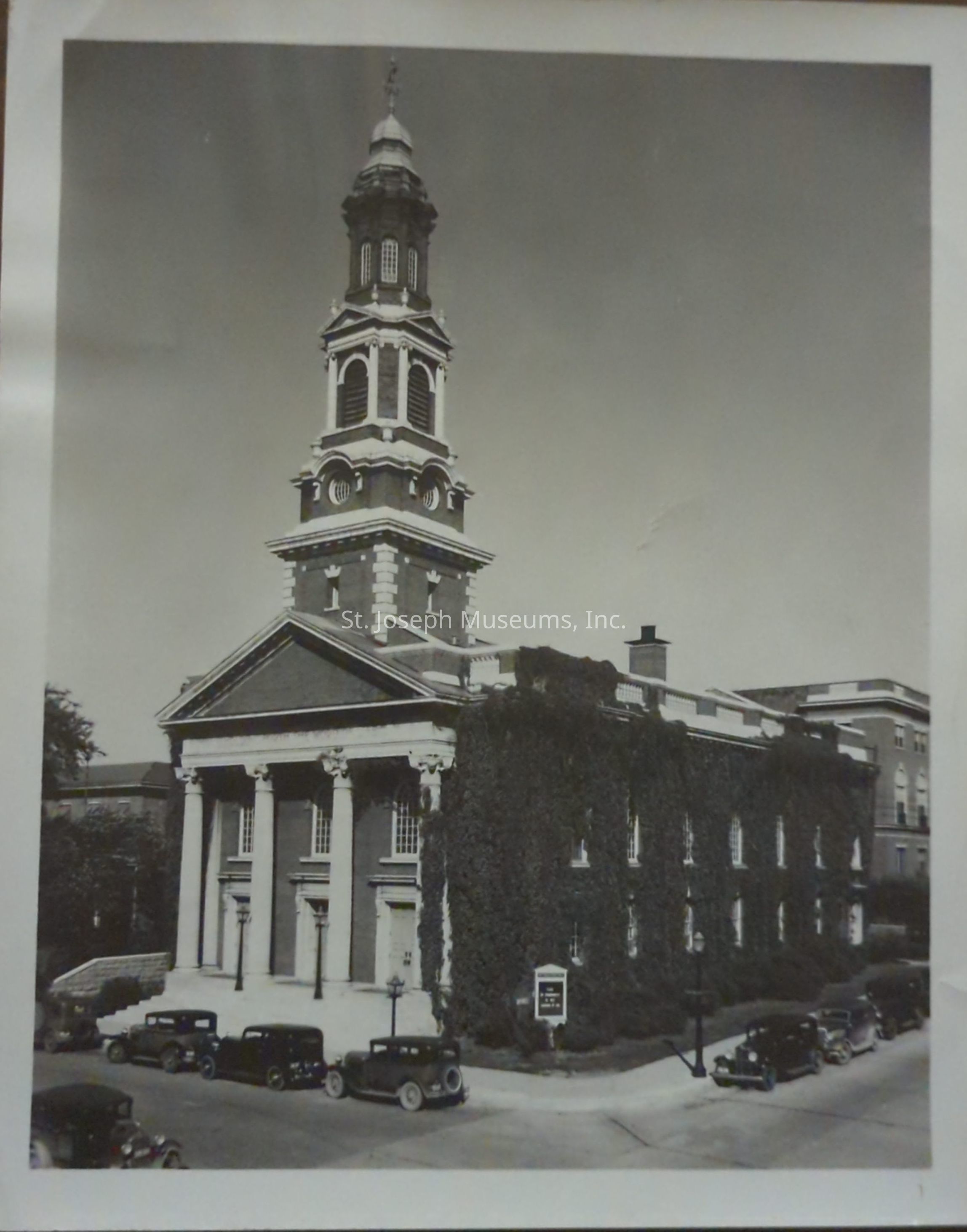 Black-and-white photograph depicting the 1st Presbyterian Church at 7th and Jules in St. Joseph, Missouri, featuring an impressive tower and classical architecture with large columns at the entrance. The building is covered in lush ivy, adding to its historic charm, and vintage cars are parked along the street.