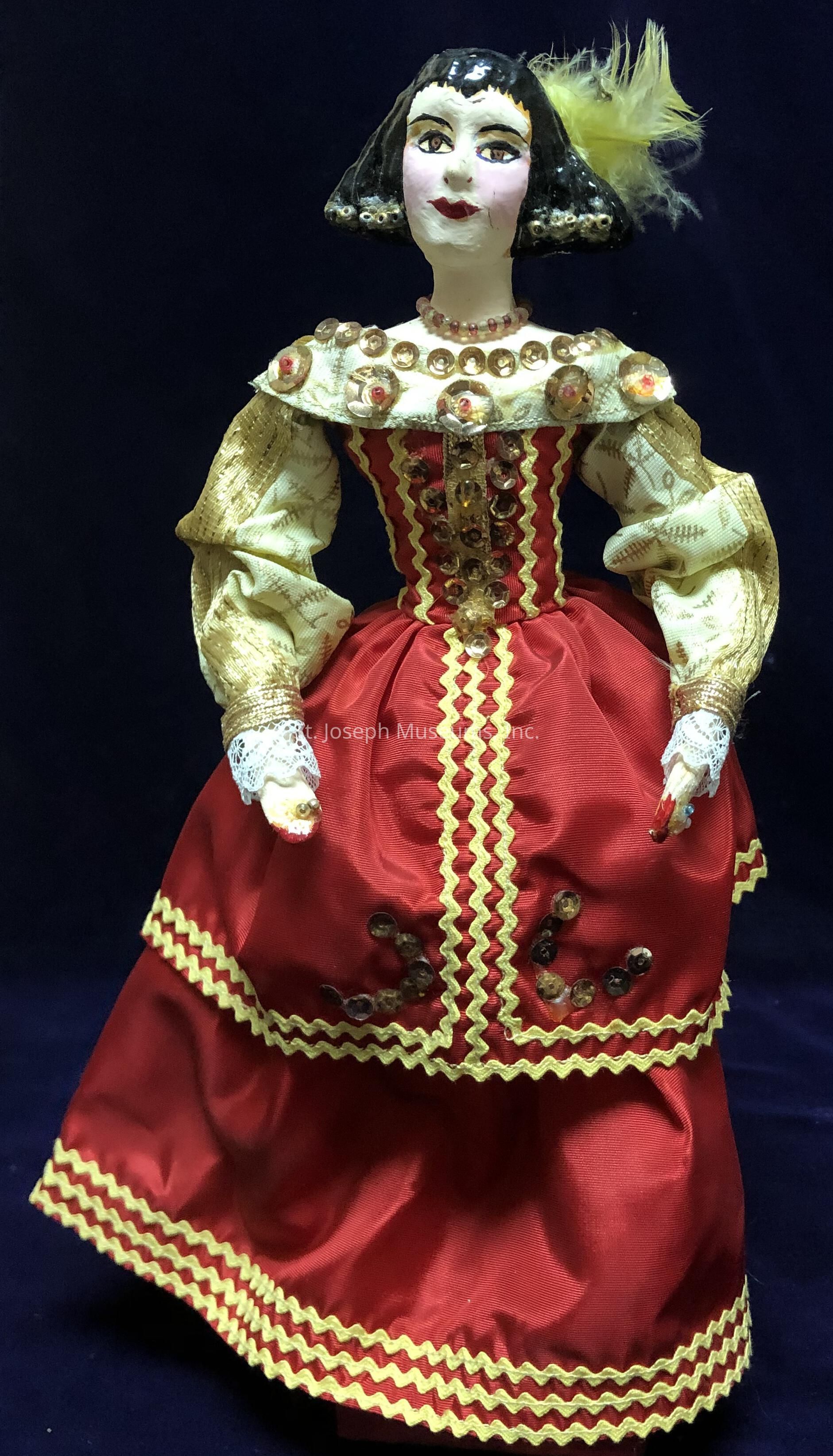The image showcases a beautifully crafted doll wearing an elaborate costume that appears to be inspired by historical fashion. The doll is dressed in a vibrant red gown adorned with ruffled yellow trim and shiny embellishments. Its bodice features intricate golden embroidery and clear sequins that catch the light, providing a luxurious effect. The puffed sleeves are made of a patterned fabric, complementing the richness of the ensemble. Additionally, the doll has a striking face with painted features including vivid eyes, a subtle smile, and a yellow feather accentuating its black, chin-length hairstyle. This arrangement, coupled with the doll's delicate pose, sets her as a refined centerpiece against a dark blue backdrop, highlighting the intricate craftsmanship and historical inspiration of the piece, evoking a sense of elegance and nostalgia reminiscent of a bygone era.