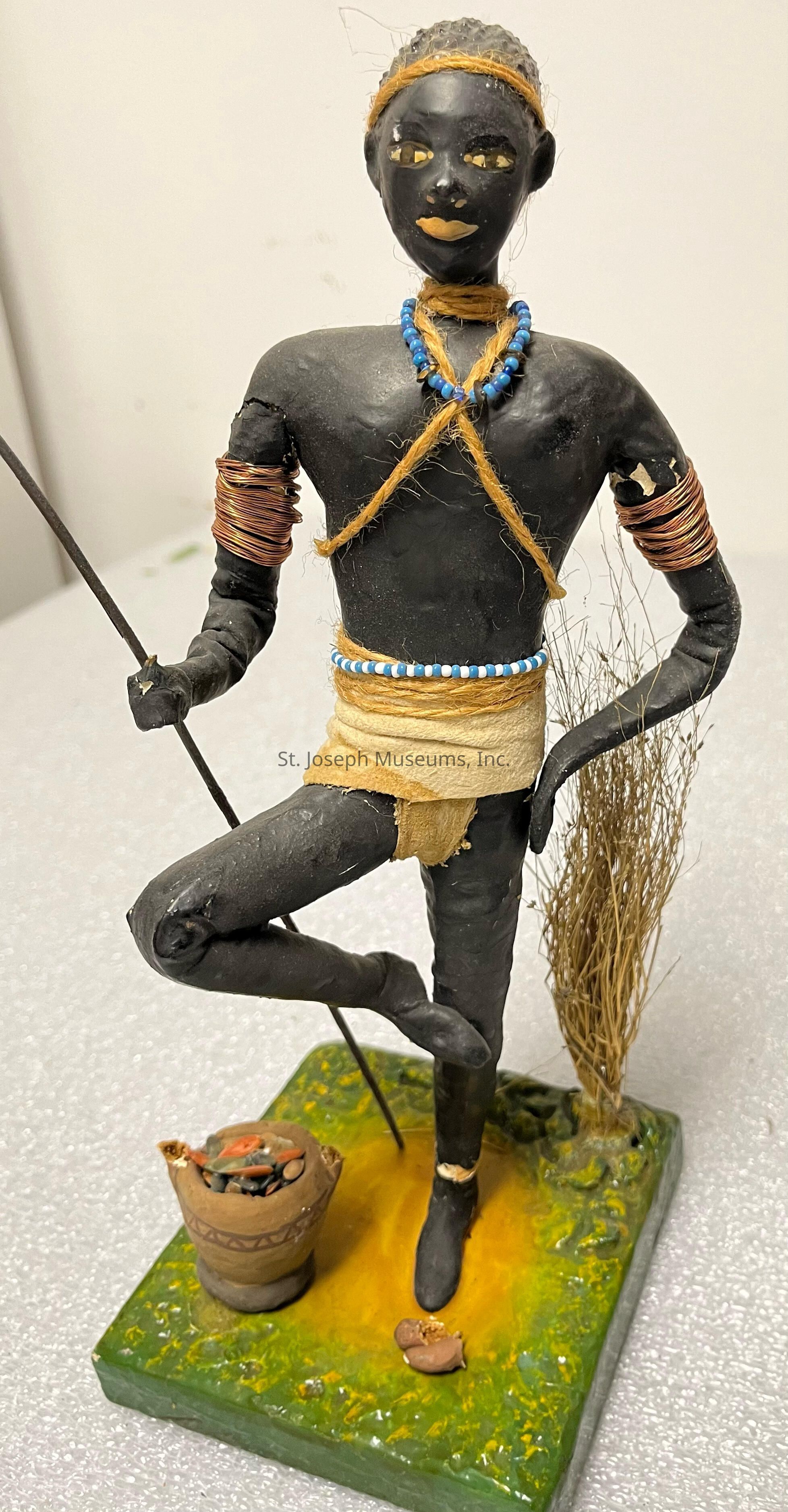 The image depicts a handcrafted figurine standing on one leg atop a textured base that resembles grass or foliage, with a small basket placed beside it. The figure is made of dark material and features intricate details, including facial features painted in gold and yellow. It is adorned with a blue bead necklace, golden headband, and a crisscross of golden twine across the torso. The arms and waist are wrapped in thin copper wire, suggesting decorative armlets. The figure’s left hand rests on its hip, while the right hand holds a slender stick or staff. Behind the figure, there are tufts of brown material representing dried grass, adding a naturalistic touch to the scene.
