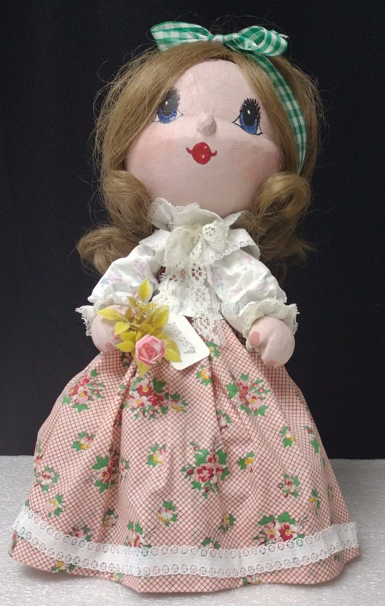 The image depicts a charming, handmade cloth doll with blue eyes and bright red lips, wearing a vintage-style dress with a floral pattern and lace details; her hair is styled in soft curls adorned with a green gingham ribbon, while she holds a small bouquet featuring a pink rose, all set against a plain black backdrop that accentuates her quaint and nostalgic appearance.