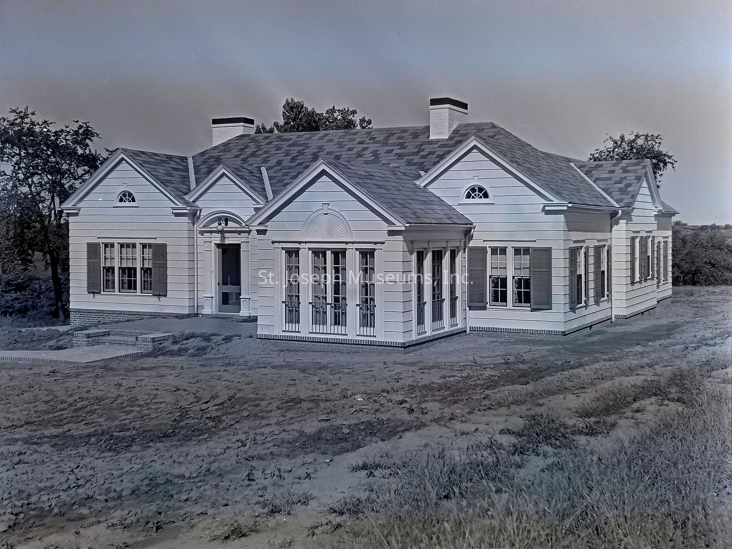 Black-and-white image showcasing the exterior of the McCord home located at 2801 Lovers Lane, St. Joseph, Missouri featuring a charming, single-story white house with a symmetrical design, multiple gabled roofs, and large windows with shutters. The property is surrounded by a natural landscape with trees and open land.