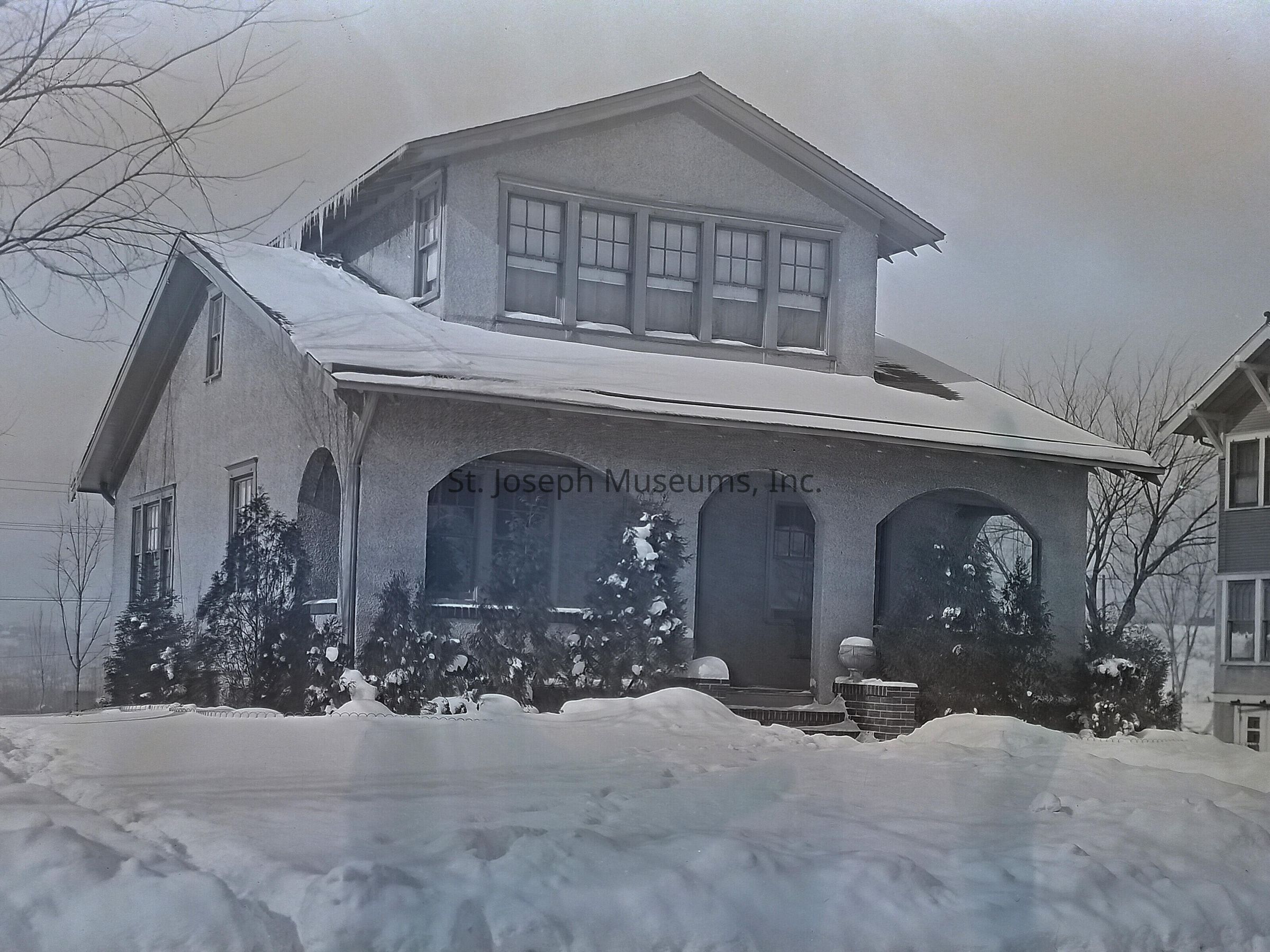 A black-and-white image depicting a snow-covered, two-story house with a distinctive front porch featuring arched openings, nestled among evergreen shrubs. The house is blanketed in snow, with icicles hanging from the roof, highlighting its wintery setting. Adjacent to the home, another building is partially visible. This scene might be from the address 2024 Lovers Lane, in St. Joseph, Missouri, based on the context provided by the envelope in which the photo was housed.