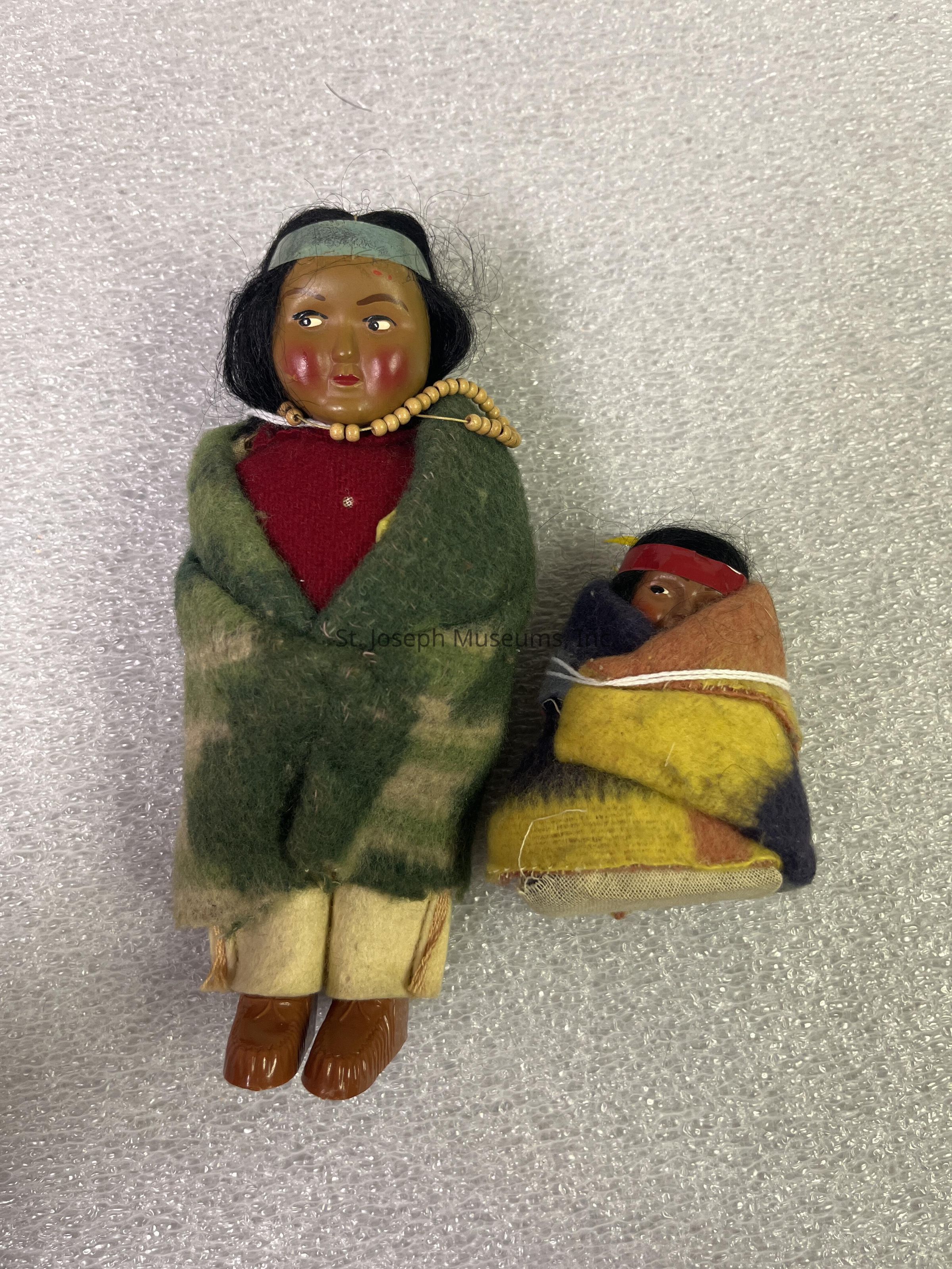 The image depicts two small dolls crafted with intricate detailing and dressed in traditional clothing. The larger doll is adorned with a green and red outfit, complemented by a bead necklace and a headband, while its facial features include rosy cheeks and black hair. It stands next to a smaller doll, which is swaddled in a colorful blanket featuring hues of yellow, red, and dark blue. The smaller doll's head peeks out of the wrap, also donning a headband and showcasing similar rosy cheeks and dark hair. Both dolls are positioned on a textured, light-colored fabric that enhances their vibrant attire and detailed craftsmanship.