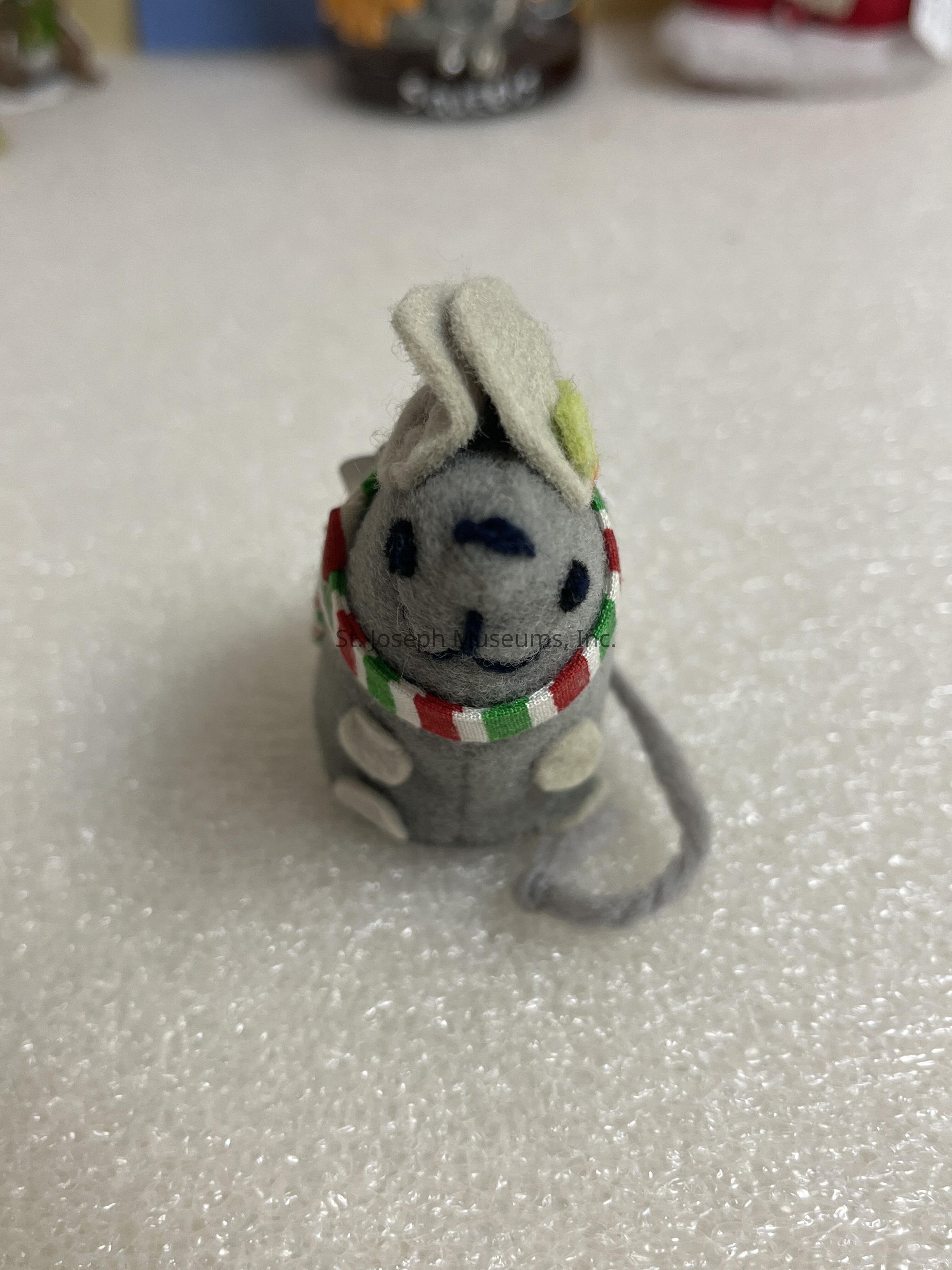 The image shows a small, cute plush toy that resembles a gray mouse with dark blue eyes and nose, sitting upright on a textured white surface. It is adorned with a colorful striped scarf in red, green, and white, adding a festive touch to its appearance. The toy features small, round, light-colored ears and a looped tail that curls beside its body, contributing to its charming and playful demeanor. The background has blurred elements, hinting at other objects, possibly decorative items, in a room setting, while the focus remains on the plush mouse, emphasizing its delightful details and crafted whimsy.