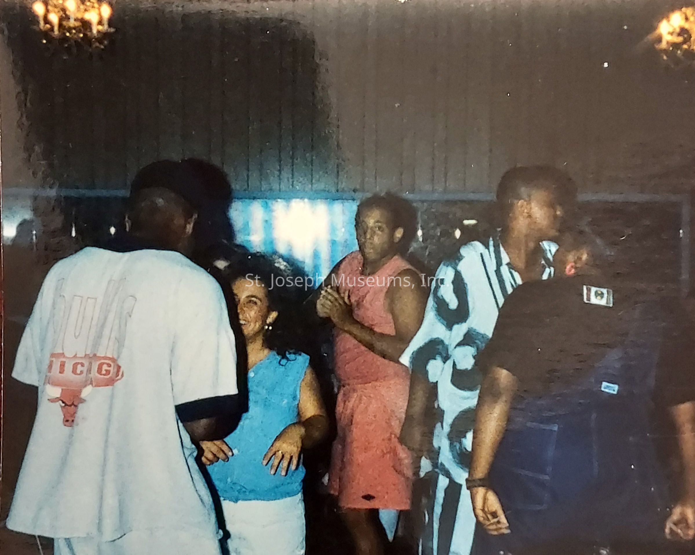 In the image, a group of people is gathered together in a dimly lit room, possibly a social event or casual gathering. One individual, who is facing away from the camera, is wearing a light grey t-shirt with "Bulls" written on it, and appears to be engaged in conversation with a woman wearing a blue sleeveless top, who is smiling warmly. To the right, another person in a pink sleeveless outfit is clapping or holding their hands together, gazing towards the camera or someone nearby. Behind them, an individual is wearing a distinctive black and white patterned shirt. The setting is informal, and there are decorative lights visible at the top, suggesting a cozy atmosphere.