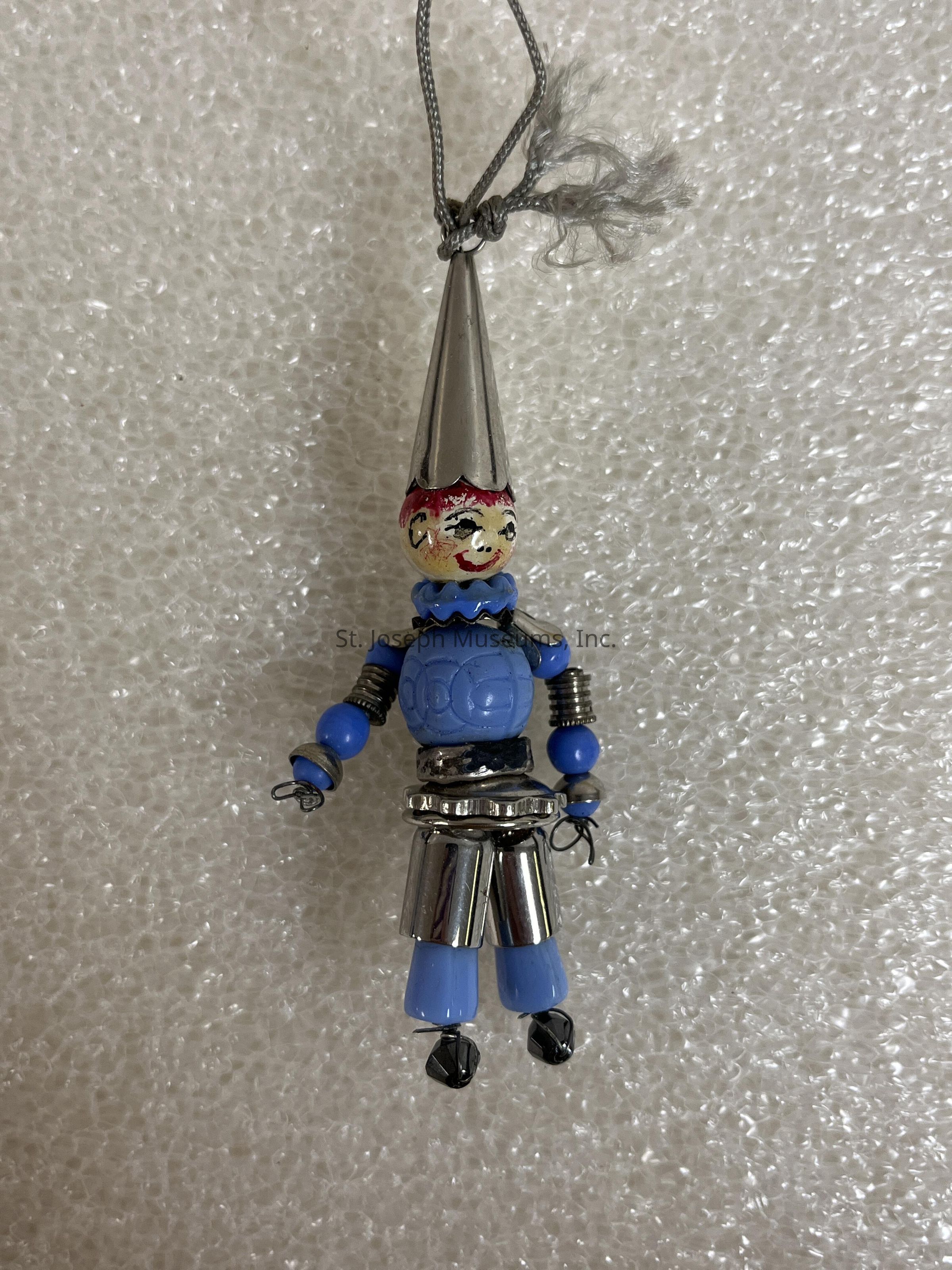 The image shows a whimsical and colorful clown ornament, featuring a painted face with a cheerful expression and bright red hair, highlighted by a tall, metallic cone-shaped hat. Its body is composed of various materials, including a blue and silver beaded torso, with playful details like metallic springs and blue limbs, giving it a toy-like appearance. This delightful figure hangs from a looped string, and the background consists of a textured, bubble wrap-like surface, which contrasts against the shiny, vibrant elements of the clown. This ornament combines elements of craft and creativity, evoking a sense of playfulness and nostalgia.