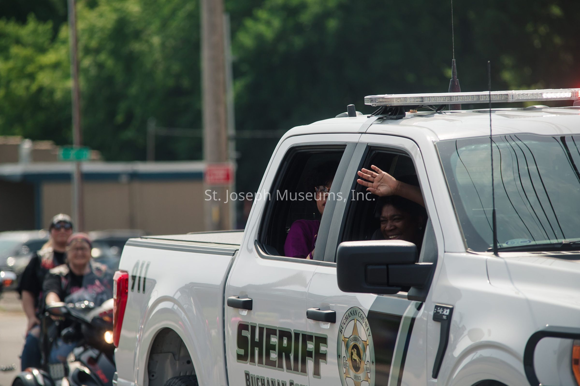 In the image, a white pickup truck marked with "Sheriff" on the side, associated with Buchanan County, is pictured driving along a street. The truck's cabin window is open, revealing a joyful individual smiling and waving to the people outside, capturing a moment of friendly interaction during what appears to be a public event or parade. In the background, blurred but still noticeable, are several people, some of whom are riding on motorcycles, contributing to the lively atmosphere of the scene. The surrounding environment features a green, tree-lined backdrop, along with urban elements such as utility poles and buildings, highlighting the blending of nature and small-town ambiance. The overall mood of the image is warm and communal, inviting a sense of togetherness and community spirit.
