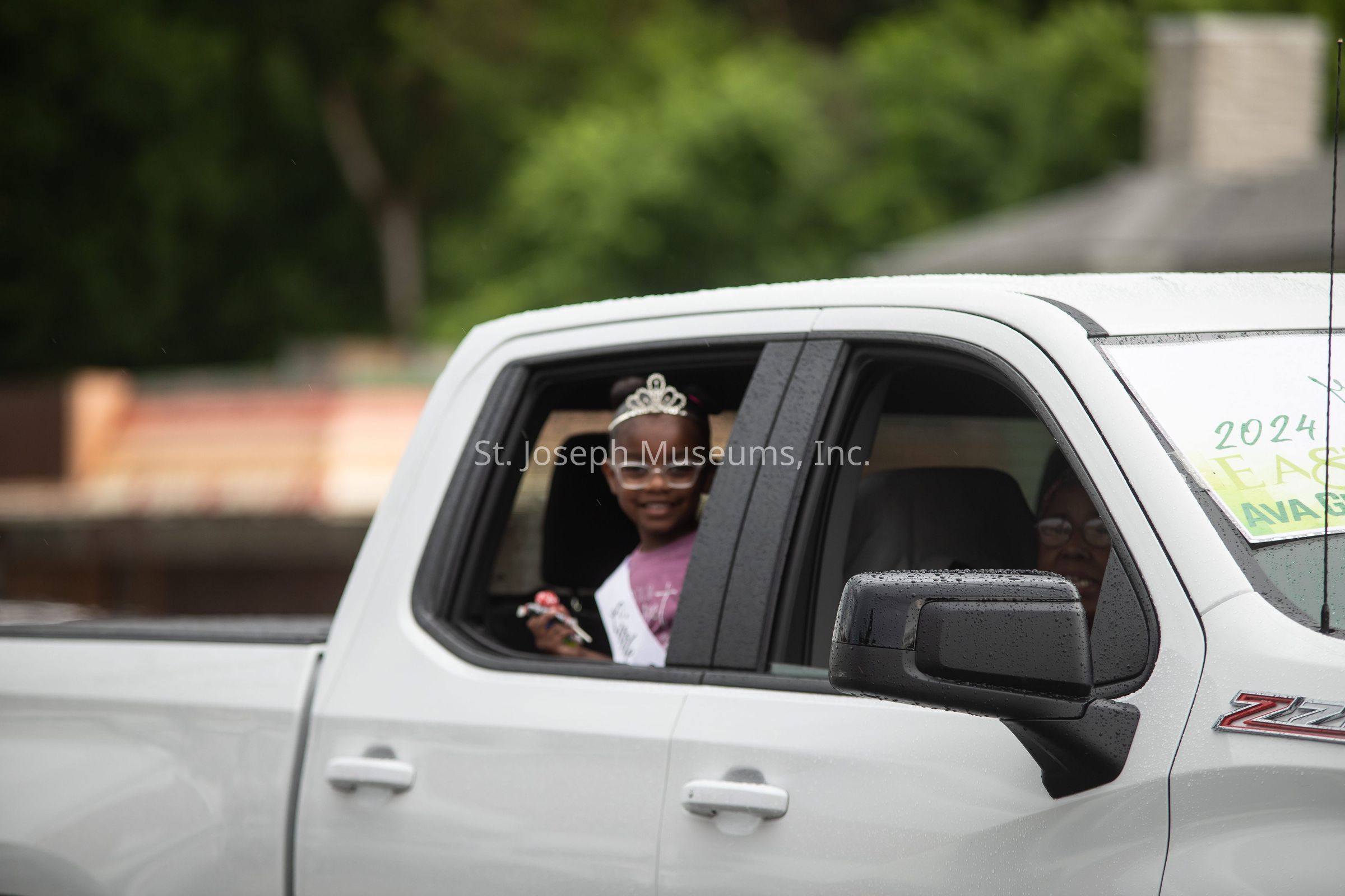 The image features a young girl smiling from the back seat of a white pickup truck. She is adorned with a tiara and wears glasses, holding a small trophy with her left hand. A sash drapes over her shoulder, indicating a celebratory event. The truck’s rear window is slightly open, and a person's face is partially visible in the driver's seat. Raindrops can be seen on the truck's surface, hinting at recent or ongoing rain. A partially visible sign on the truck's dashboard reads "2024" alongside some text, suggesting a possible school or community event such as a graduation or parade. The blurred background consists of greenery and a building, adding to the community setting.
