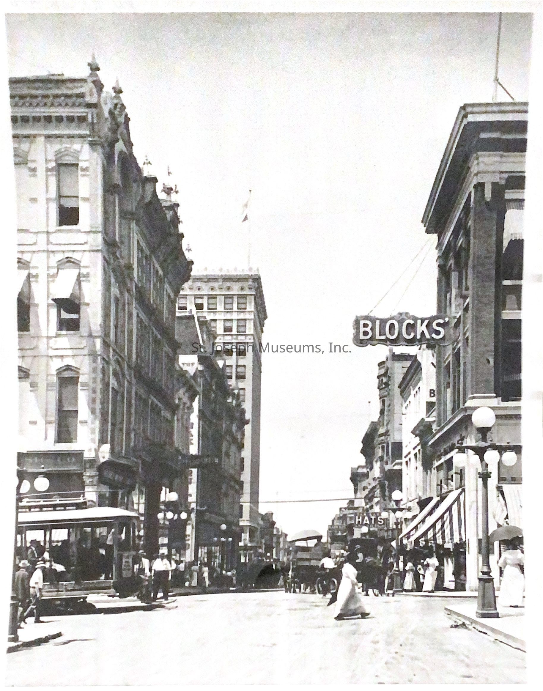 Black-and-white photo capturing a bustling street scene at the corner of 6th and Felix, looking west. Buildings with intricate architectural details line both sides, while a streetcar and pedestrians fill the scene. People dressed in period attire walk along the sidewalks as horse-drawn carriages move down the street.