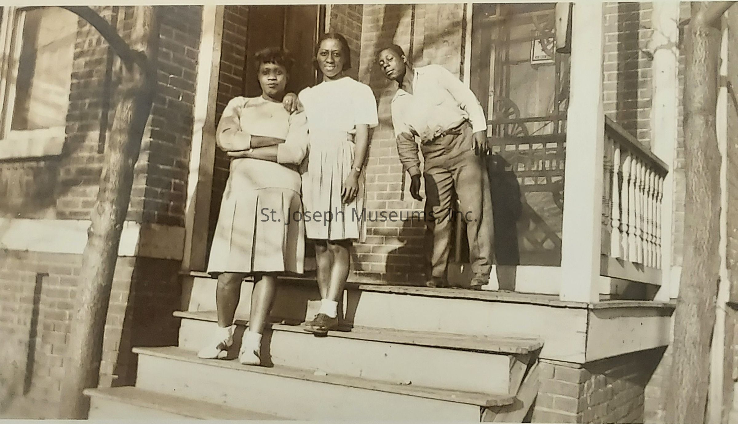 The image depicts three individuals standing on the steps of a brick building. The person on the left is a woman wearing a light-colored, knee-length skirt, with her arms crossed and standing confidently. In the center is another woman wearing a dress with pleats at the skirt's bottom, standing with a gentle smile. On the right is a man leaning casually, dressed in a shirt and pants, with one hand in his pocket. The wooden porch and visible railing contribute to the warm and relaxed atmosphere, and sunlight casts shadows on the wall, suggesting a pleasant day. The scene exudes a timeless quality, capturing a moment of camaraderie and ease among the three individuals in a domestic setting, highlighting the everyday simplicity and connection among them. The elements of the photograph—clothing style, architecture, and the sepia tone—suggest it is from a past era, giving it a nostalgic and historical feel. The composition is balanced, and their expressions hint at a shared moment of joy or perhaps a sense