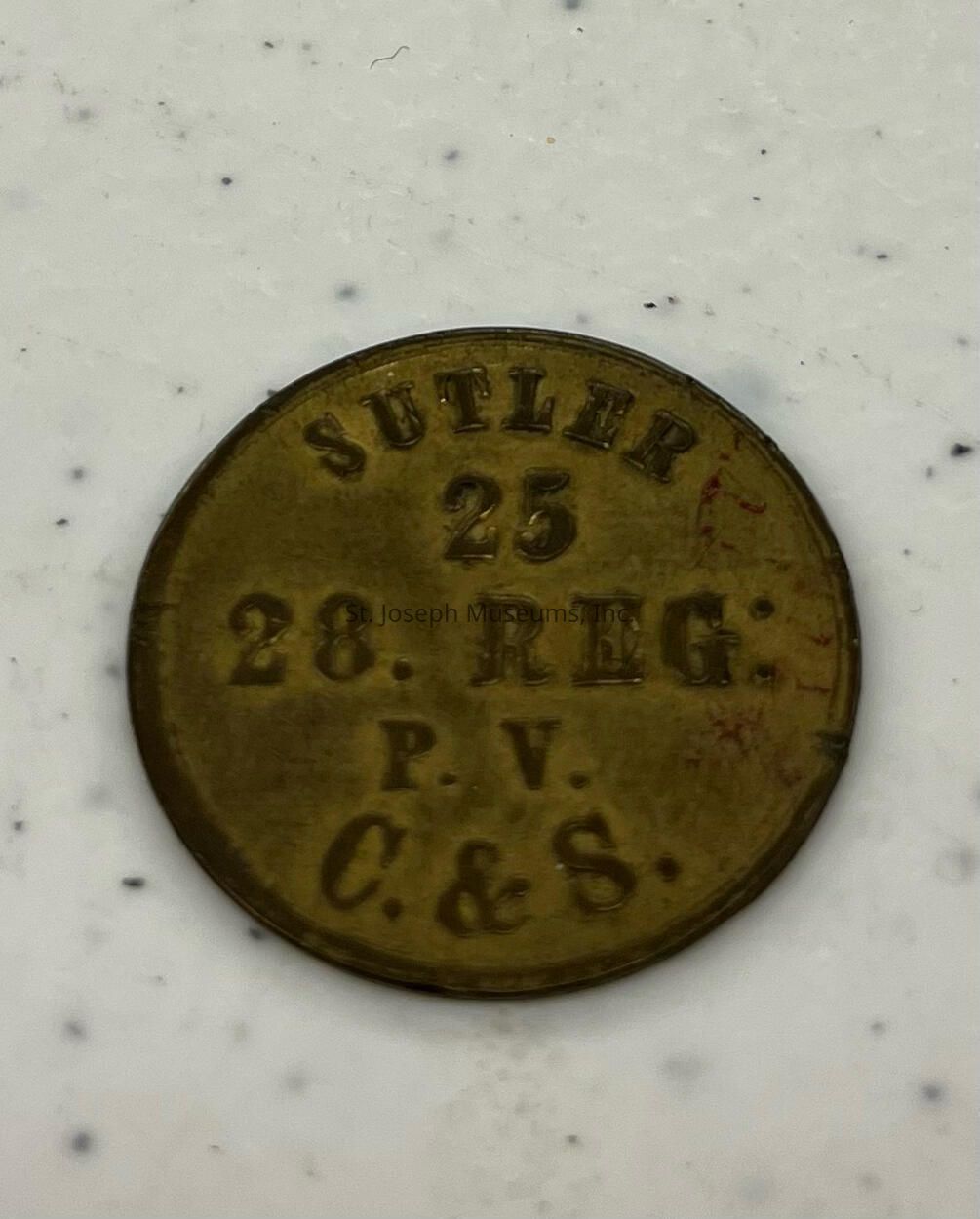 The image features a round brass token with engraved text. The top of the token is inscribed with "SUTLER," followed by the number "25" beneath it. Below this, the text reads "28. REG:" indicating possibly a regiment designation. Further down, the letters "P. V." are visible, and at the bottom, it reads "C. & S." The token appears to be well-worn, hinting at its age, and sits on a light background speckled with small black dots, which provides contrast to the token’s metallic color and highlights its historical significance.