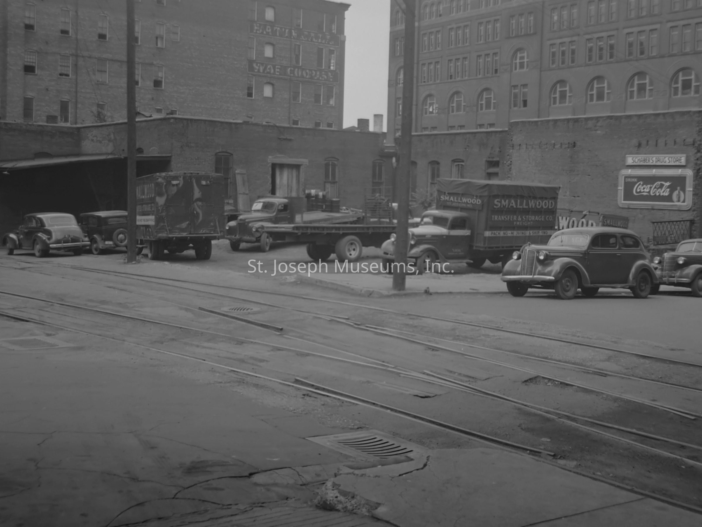 Black-and-white image depicts the rear view of the Smallwood Transfer & Storage Co. building and its adjacent parking area in St. Joseph, Missouri, showcasing vehicles parked alongside the structure. The brick façade is visible, with advertising signs in the background, including a Coca-Cola and a Schreiber's Drug Store sign.