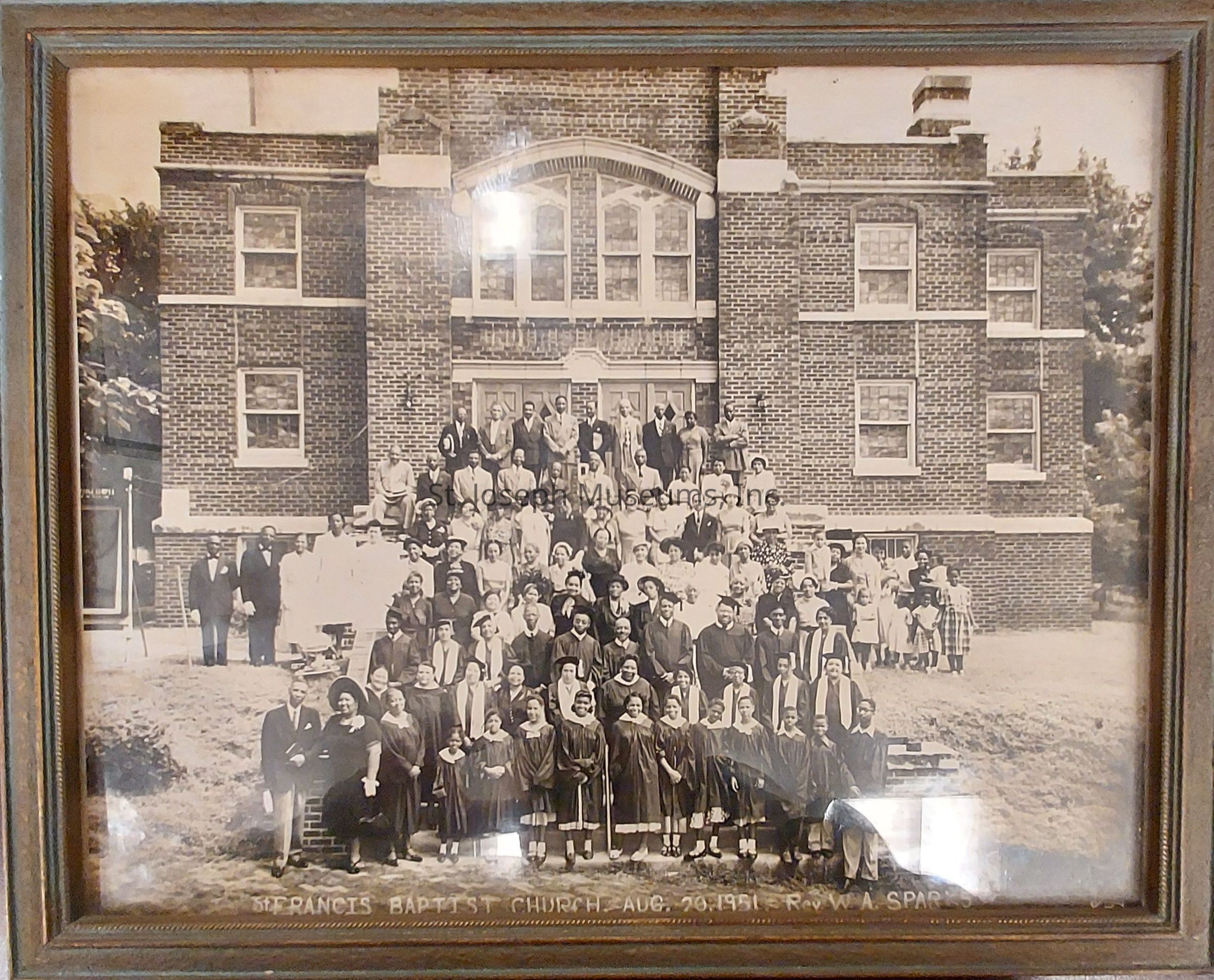 The image depicts a large group of people assembled in front of a brick building identified as "St. Francis Baptist Church" according to the text at the bottom. The photograph, dated August 26, 1951, includes men, women, and children dressed in formal attire, with some individuals seated and others standing on the steps leading up to the church entryway. The building features several windows, and the people are arranged in a pyramid-like formation that ascends toward the entrance. Lush trees are visible on either side of the structure, framing the scene with a natural backdrop.