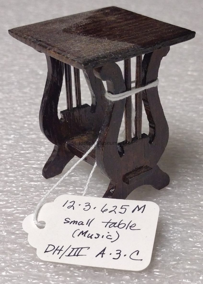 The image features a small, intricately designed table resembling a piece of furniture, possibly a miniaturized model, with a dark wooden finish and a flat, rectangular top supported by elegantly curved legs that give the appearance of ornate craftsmanship. This miniature table is described in a handwritten label attached with white string, which reads "small table (Music)" along with other classification codes. The surface on which the table rests appears to be a textured, protective material, such as bubble wrap, highlighting the careful handling and preservation of the item.