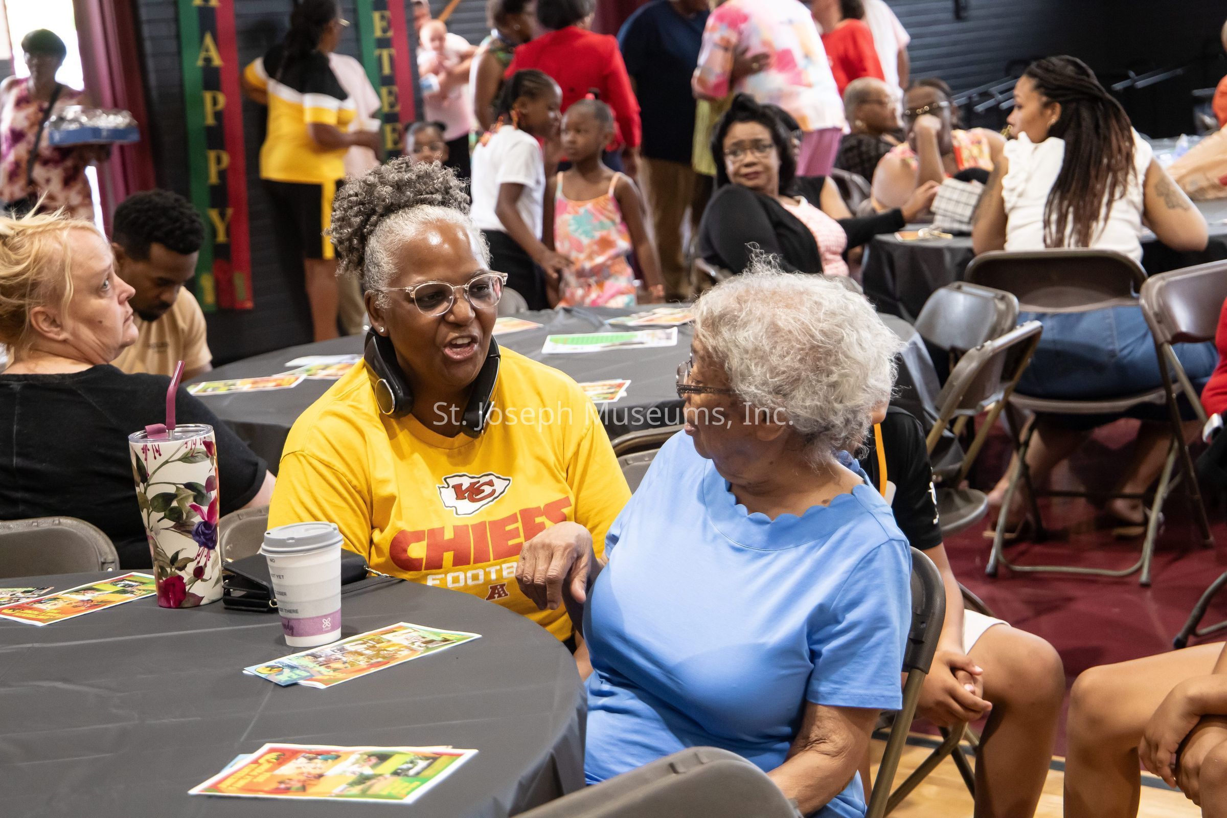 The image depicts a lively social gathering, where people of various ages are seated around tables covered with black tablecloths. In the foreground, two women are engaged in a friendly conversation, one sporting a yellow "Chiefs Football" shirt and headphones around her neck, while the other wears a blue blouse, and they appear to be enjoying each other’s company. Around them, other attendees are seated or standing in the background, creating a bustling atmosphere with interactions among small groups. The room is decorated with colorful banners and there are drinks and cards placed on the tables, suggesting a celebratory or community event where people are interacting warmly.