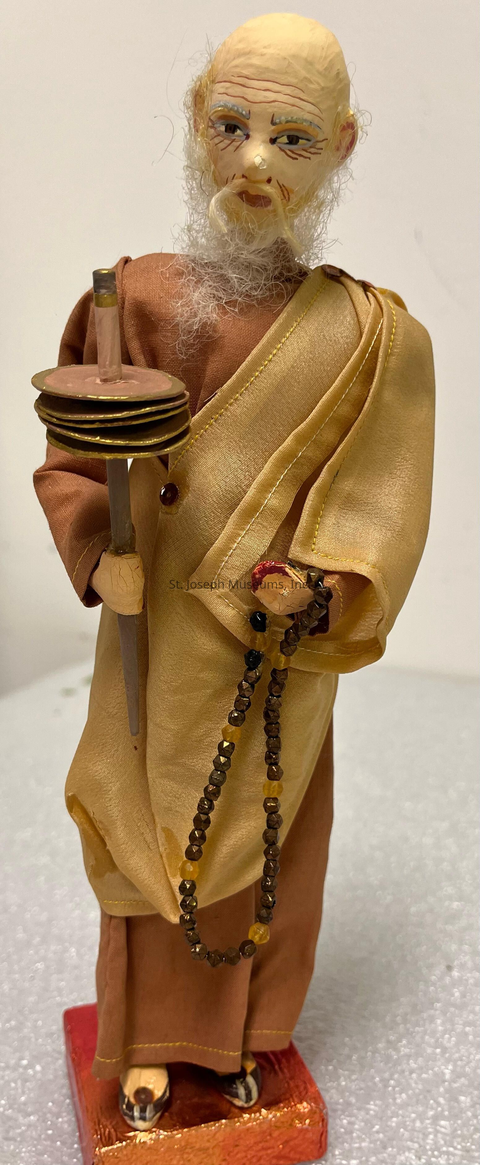 The image depicts a handcrafted figurine of an elderly man, designed with intricate details. He has a white beard and wrinkled facial features, embodying wisdom and age. The figure is dressed in robes, combining shades of brown and golden yellow, with a wrap draped elegantly over one shoulder. In his left hand, he carries what appears to be circular objects, possibly books, stacked vertically on a stick, while his right hand holds a string of brown beads, suggesting spirituality or meditation practices. The figure stands on a red rectangular base, and his feet appear in traditional footwear. This artistic piece captures a serene, contemplative expression, complemented by a textured background that adds depth and contrast to the figure.
