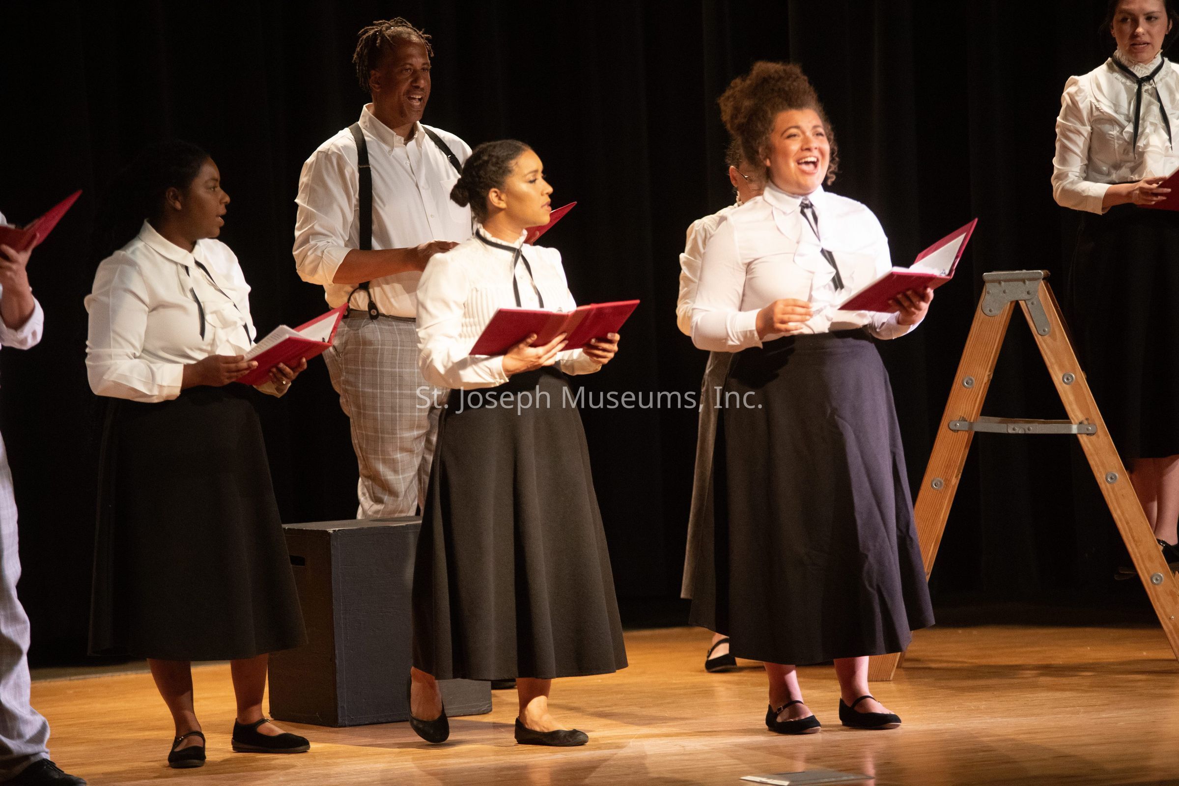 In the image, a group of performers stands on a wooden stage, engaging in what appears to be a theatrical or musical performance. The individuals are predominantly dressed in coordinated black skirts and white shirts, with some wearing suspenders, creating a formal and uniform look. They hold red folders, likely containing scripts or sheet music, indicating a choral or dramatic reading. One performer, positioned near the center, is smiling broadly, possibly delivering a line or singing, suggesting an atmosphere of enjoyment and engagement. To the right, a wooden ladder is visible, possibly a prop or part of the stage setup, hinting at a creative or symbolic element in the performance. The background is dark, drawing focus on the performers and enhancing the stage presence, while the warm lighting highlights the expressive and lively nature of the participants.