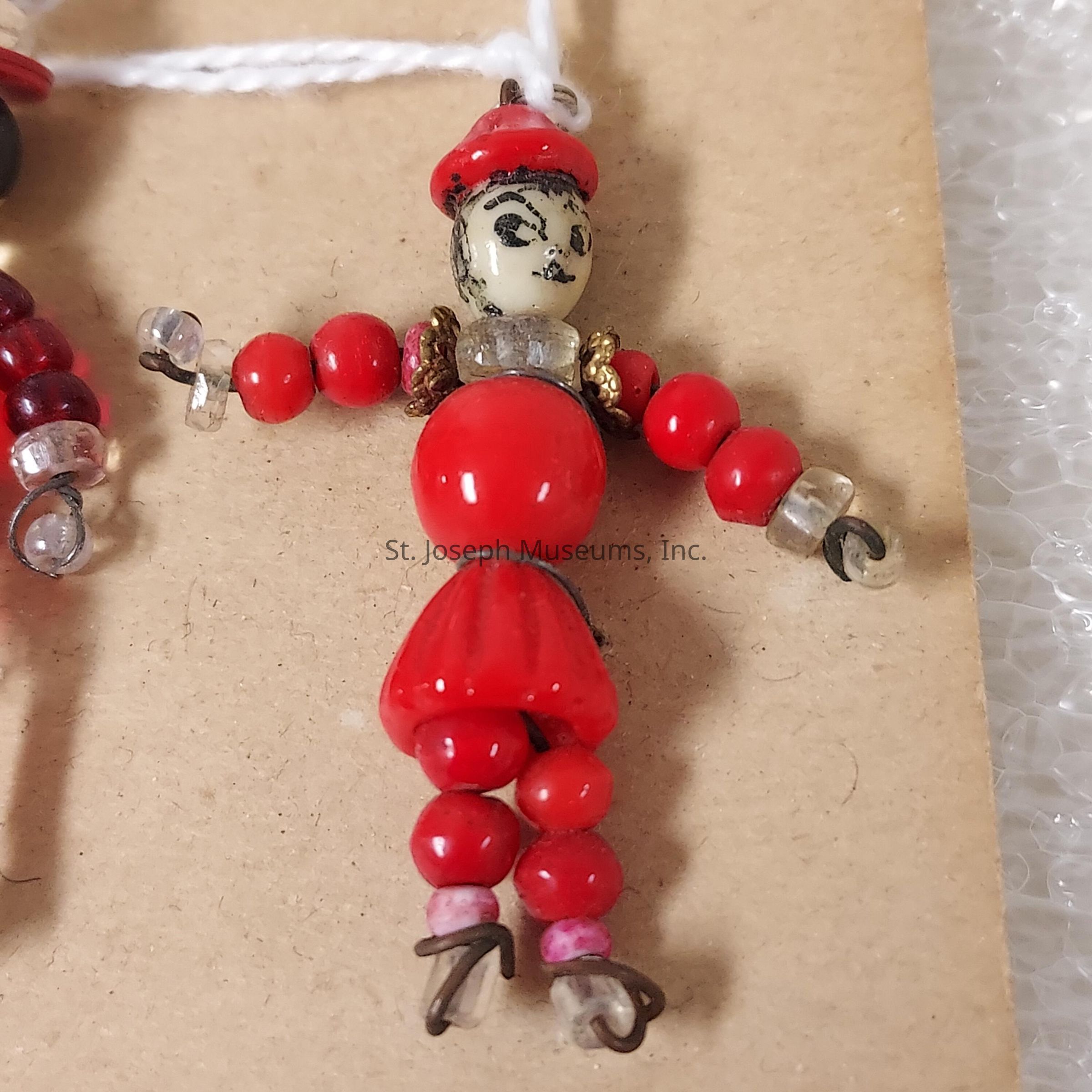 The image shows a handcrafted beaded figure, artistically composed with red and clear beads on a neutral background. The figure has a small, detailed head featuring a painted face, adorned with a red hat matching the red beads that form the body and limbs. The figure's arms and legs are articulated with alternating clear and red beads that create a charming and vibrant aesthetic. Metal loops and wires are used to connect the beads, giving the piece structure and form. The figure's charming pose and the subtle shine of the beads create a playful and artistic expression, evoking a sense of nostalgia and creativity.