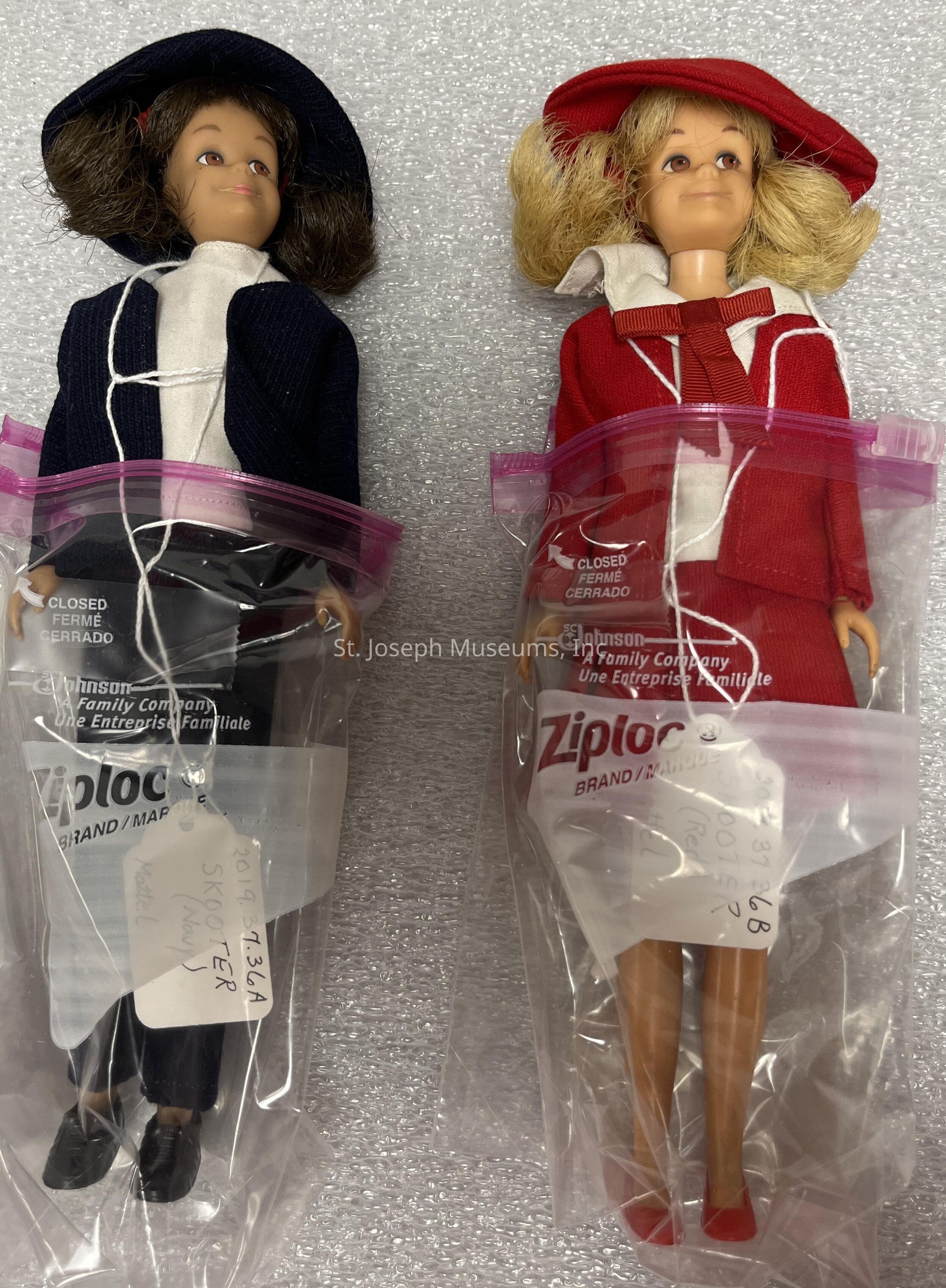 The image features two vintage dolls, each encased in individual Ziploc bags with visible tags labeled "SKOOTER." The doll on the left is dressed in a navy blue outfit with a matching hat, while the doll on the right is adorned in a red ensemble, also with a coordinating hat. Both dolls have distinctive hairstyles: the one on the left has short brown hair, and the one on the right sports blonde hair styled in curls. The Ziploc bags are sealed, ensuring preservation, and are placed on a textured surface that appears to provide cushioning. Each doll's outfit is further detailed with contrasting white accents.