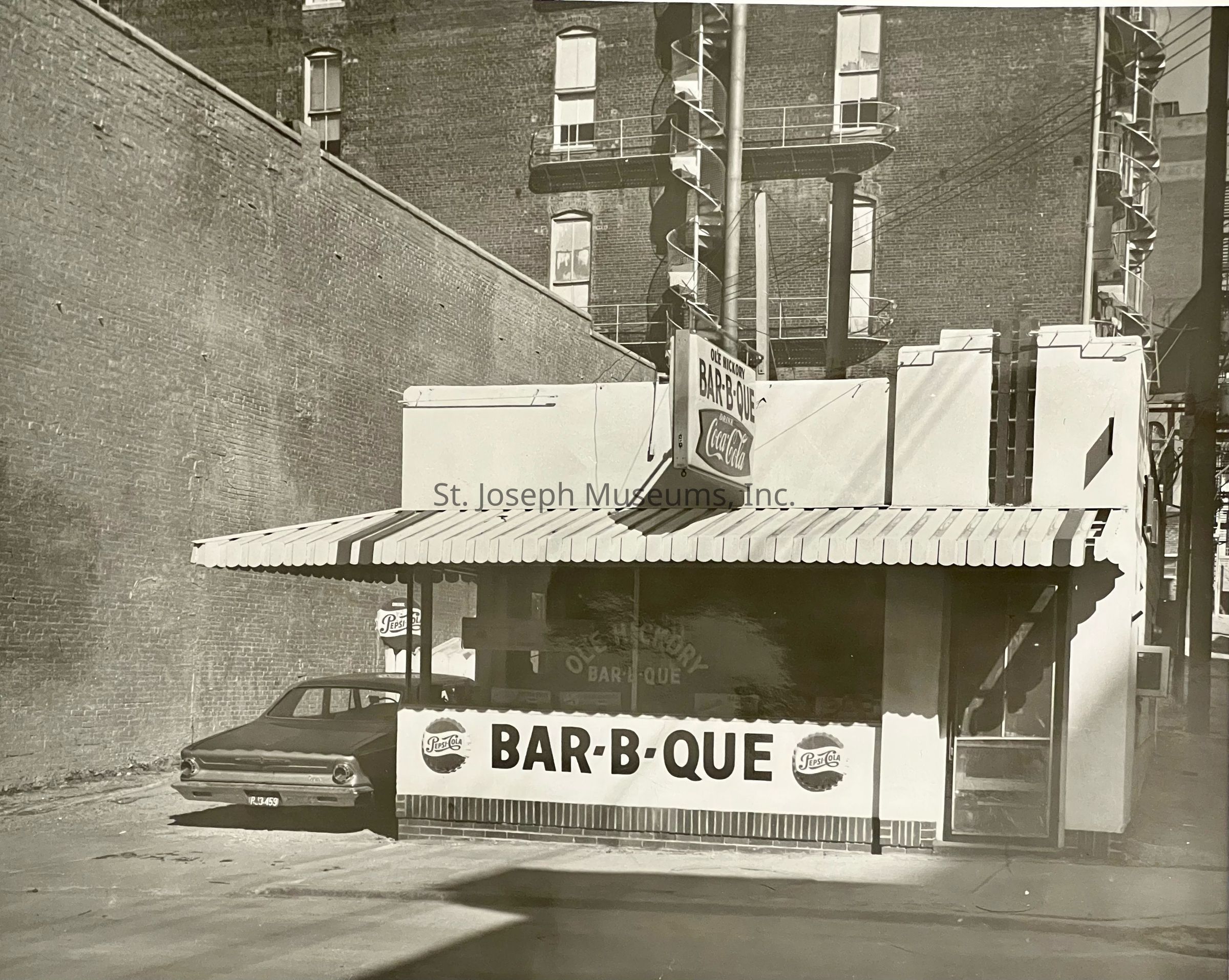 Black-and-white photograph depicting the storefront of Ole Hickory Bar-B-Q located on the north side of Felix, between 4th and 5th streets. The quaint establishment features a prominent awning with bold "BAR-B-Q" signage and a vintage Coca-Cola sign hanging above. A car is parked beside the building.