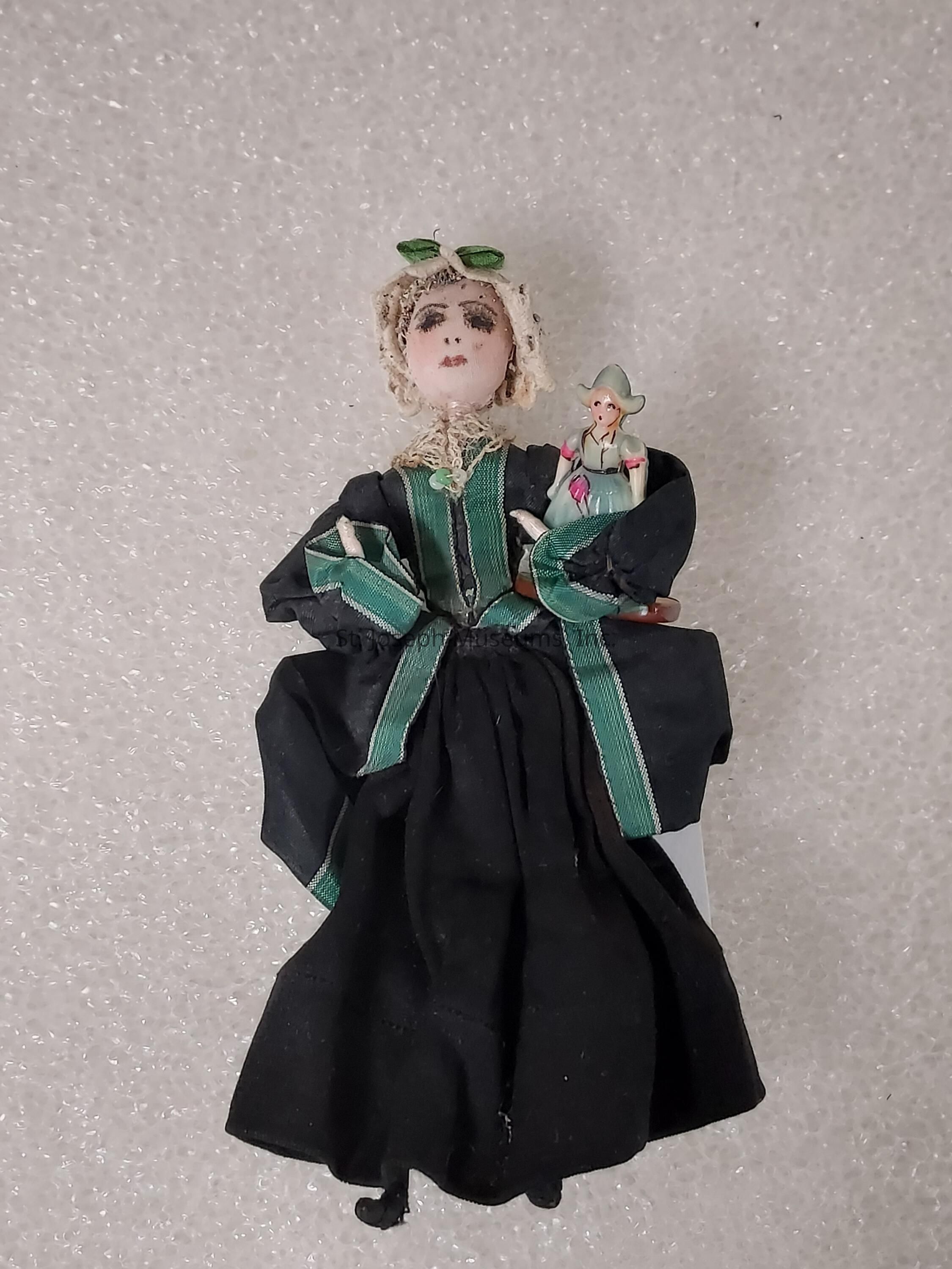 The image depicts a doll clad in a black and green dress with fine detailing, including lace and a green ribbon. The doll has a porcelain-like face with painted features and wears a lace bonnet adorned with a green bow. Resting on the doll’s arm is a smaller, intricately-detailed figure wearing a light-colored dress and hat. The backdrop is a textured, neutral surface that highlights the intricate detailing of the doll's attire and features. The overall appearance of the doll suggests a vintage or antique style, adding a sense of historical charm and elegance to the figure.