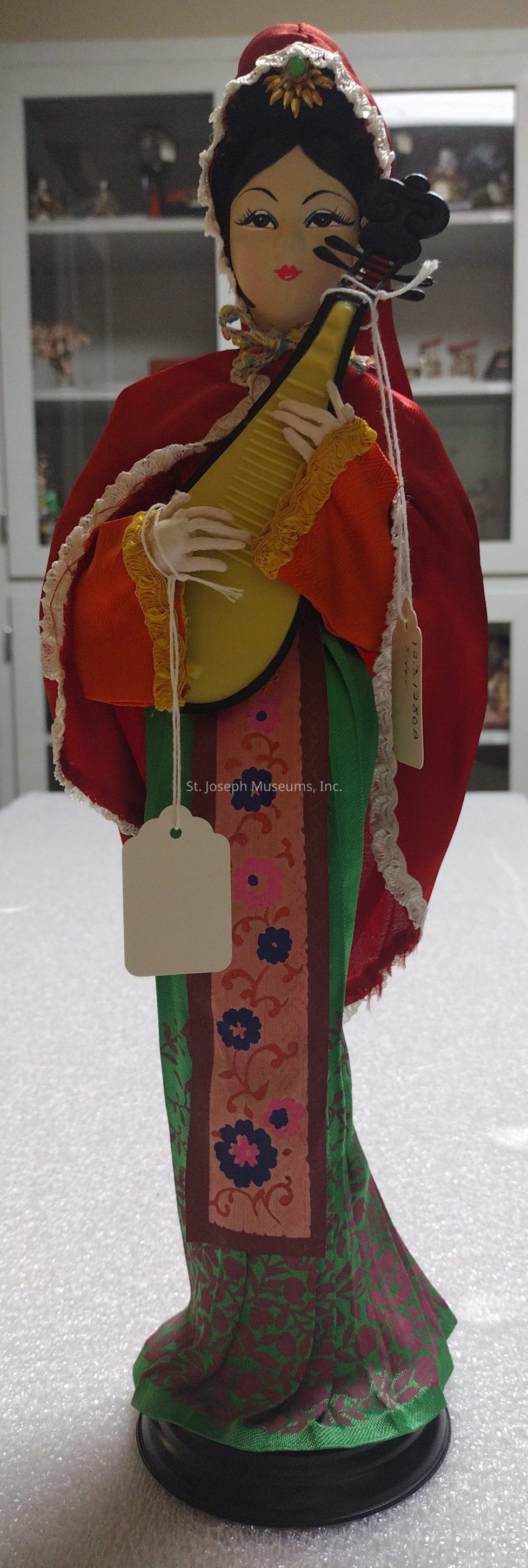 The image depicts a traditional doll dressed in vibrant and elaborate attire, likely modeled after a cultural or historical figure, with the doll holding a stringed musical instrument resembling a pipa. The doll is adorned with a red and white headdress, floral patterns on its clothing, and stands on a black circular base. Its intricate clothing, featuring red, green, and orange hues, is detailed with lace and floral embroidery, giving a sense of elegance and careful craftsmanship. Two white tags hang from the doll, suggesting it might be a collectible or display piece. The background shows some shelves and a glass cabinet.