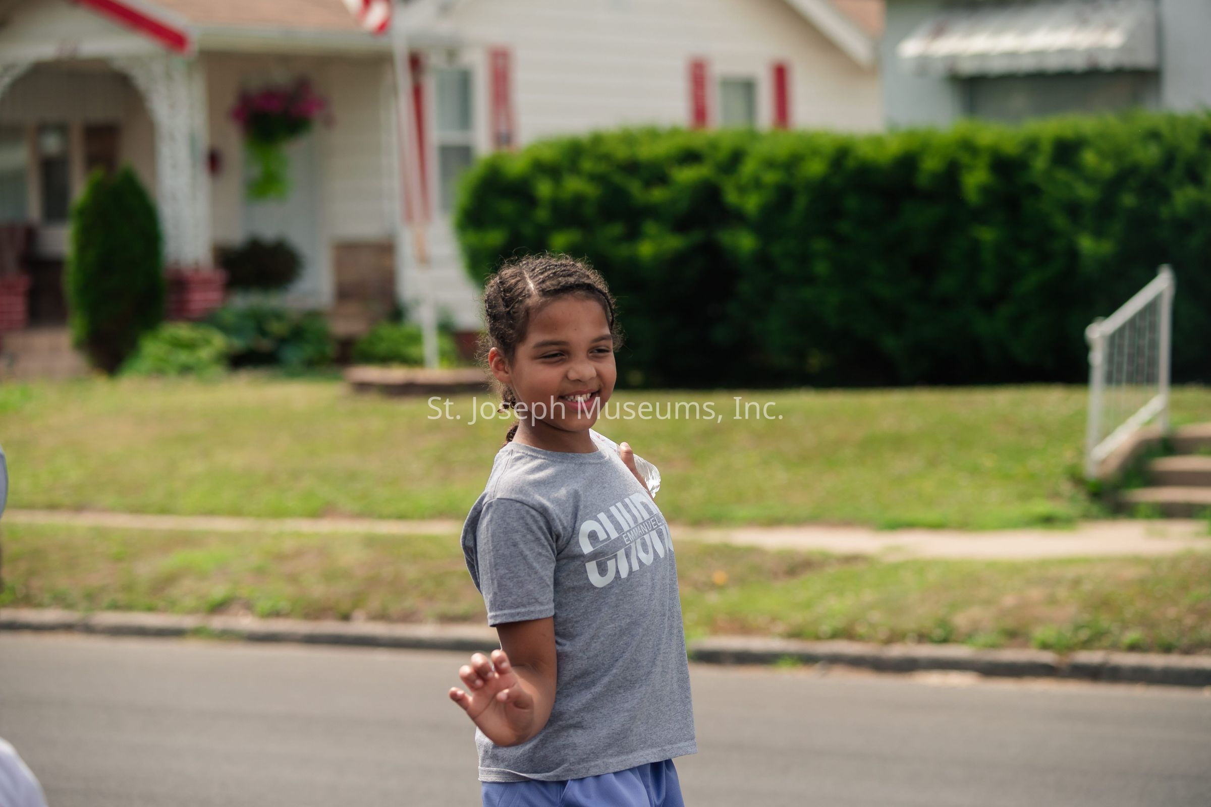 A young girl smiles joyfully while standing on a residential street, wearing a gray t-shirt and blue shorts; she has braided hair and is holding a small white object in her left hand, possibly a paper or cloth, while playfully waving with her right hand. The background shows a neatly maintained suburban environment with a white house adorned with red shutters and a front garden that is vibrant with greenery, including shrubs and well-kept lawn. An American flag is visible, suggesting it might be a patriotic holiday or event. The ambiance is cheerful and lively, capturing the essence of a sunny day filled with outdoor activities or possibly a parade or community gathering. The girl's expression and body language convey excitement and carefree enjoyment, embodying the spirit of a small-town neighborhood where community ties are strong and children can play freely in the safety of their surroundings.