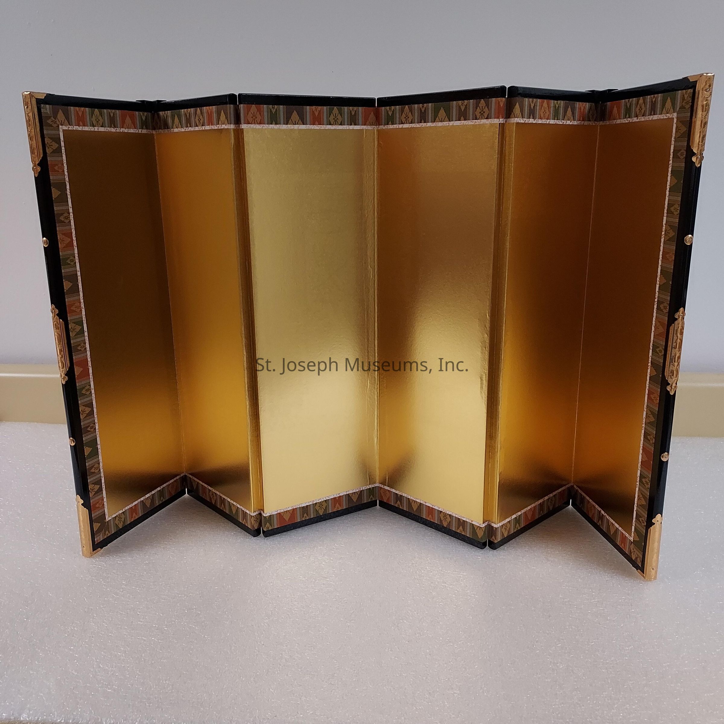 The image features a beautifully crafted folding screen with a gold-leaf finish, displaying a series of connected panels that create a striking visual impact. The screen stands elegantly on a light-colored surface, suggesting a refined and sophisticated setting. Each panel is bordered with intricate designs featuring geometric patterns in muted tones, adding a subtle yet decorative touch to the overall appearance. The edges of the screen are accentuated with dark trims, which provide a strong contrast to the shimmering golden center, highlighting the luxurious nature of the piece. This folding screen, with its reflective surface and careful attention to detail, serves not only as a functional partition but also as an ornamental element, likely intended for interior decor in an upscale environment. The golden hue radiates a warm glow, creating an inviting atmosphere, while the folding design offers versatility and flexibility in its use, making it both a practical and aesthetically pleasing addition to the space.
