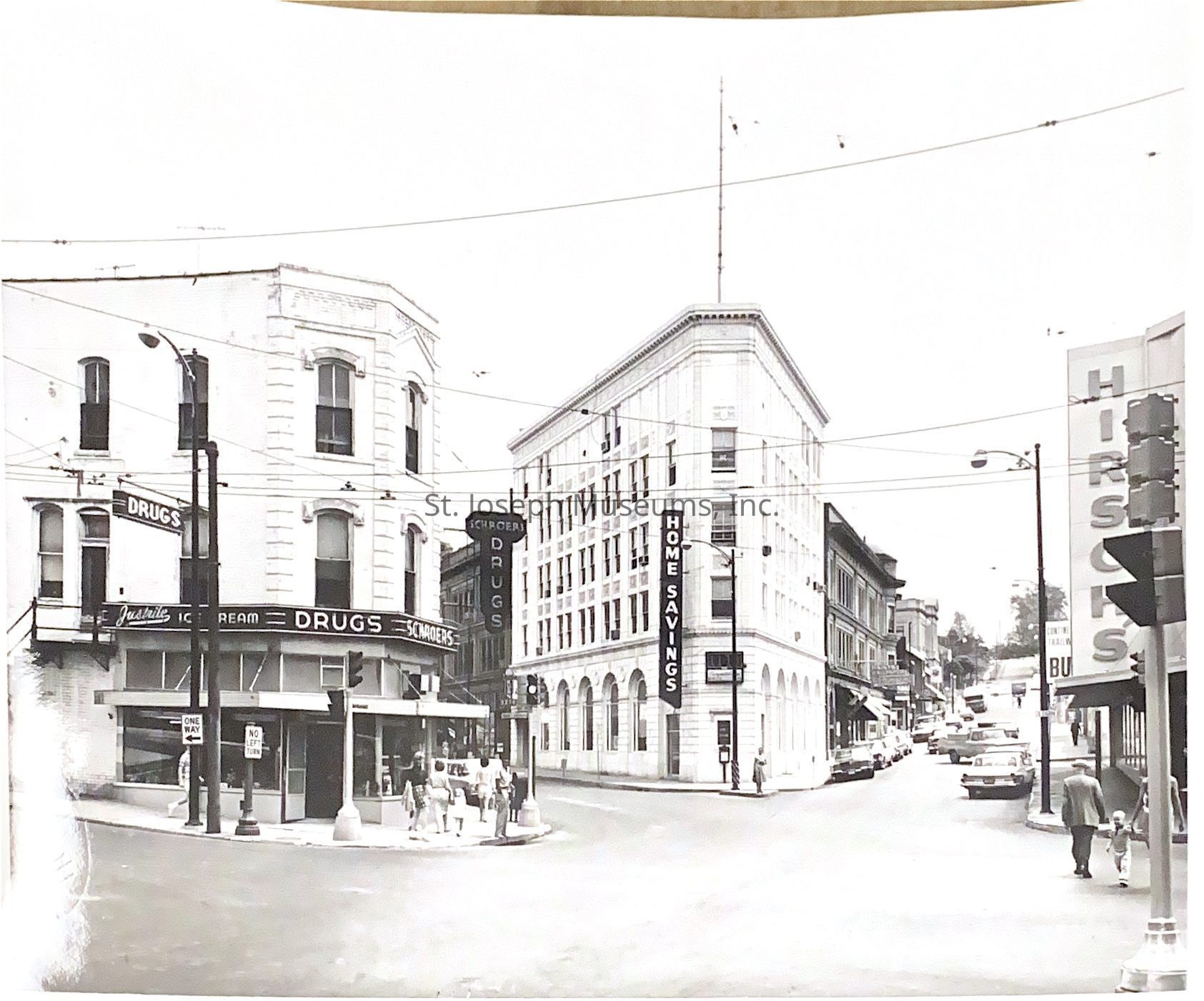 Black-and-white photograph depicting a bustling intersection featuring several prominent storefronts, including Schroers Drugstore on the left corner, characterized by its classic signage advertising pharmacy services and ice cream, and the multi-story Home Savings building in the center with bold vertical signage. To the right, part of Hirsch’s store sign is visible. The streets are busy with pedestrians crossing and vehicles parked along the sides, contributing to a lively urban scene of St. Joseph past.