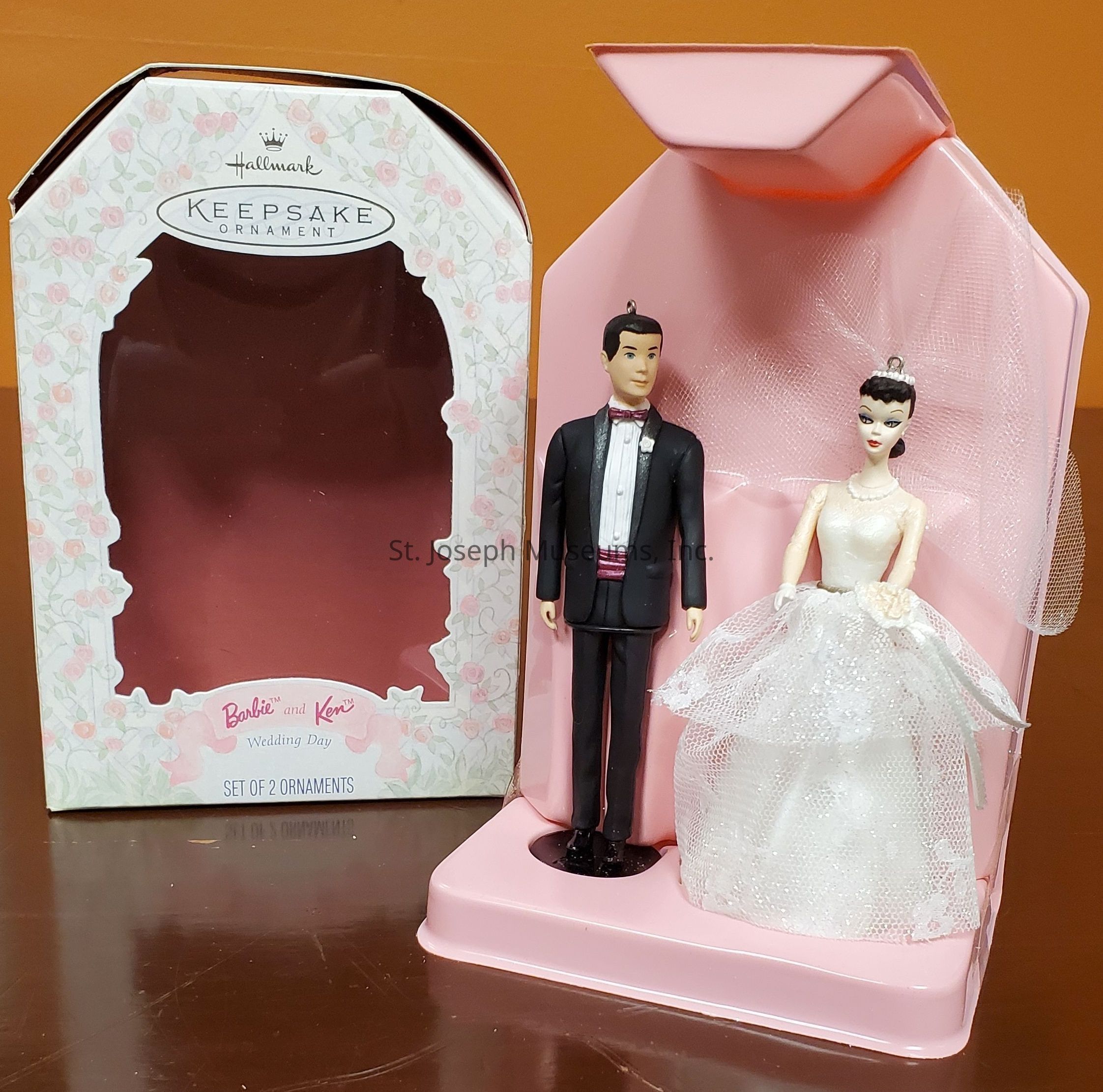 The image features a "Hallmark Keepsake Ornament" set that includes two figurines styled as a bride and groom. The groom, dressed in a smart black tuxedo with a bow tie, stands beside the bride, who is adorned in an elegant white wedding gown with intricate lace and tulle detailing. Both figurines have small loops on their heads, indicating they are intended to be used as ornaments. The set is displayed in a pink, open card-like packaging that holds the figurines securely in place, and an empty, floral-patterned box with a clear window is situated beside it, suggesting that both serve as part of a collectible wedding-themed series identified as "Barbie and Ken Wedding Day."