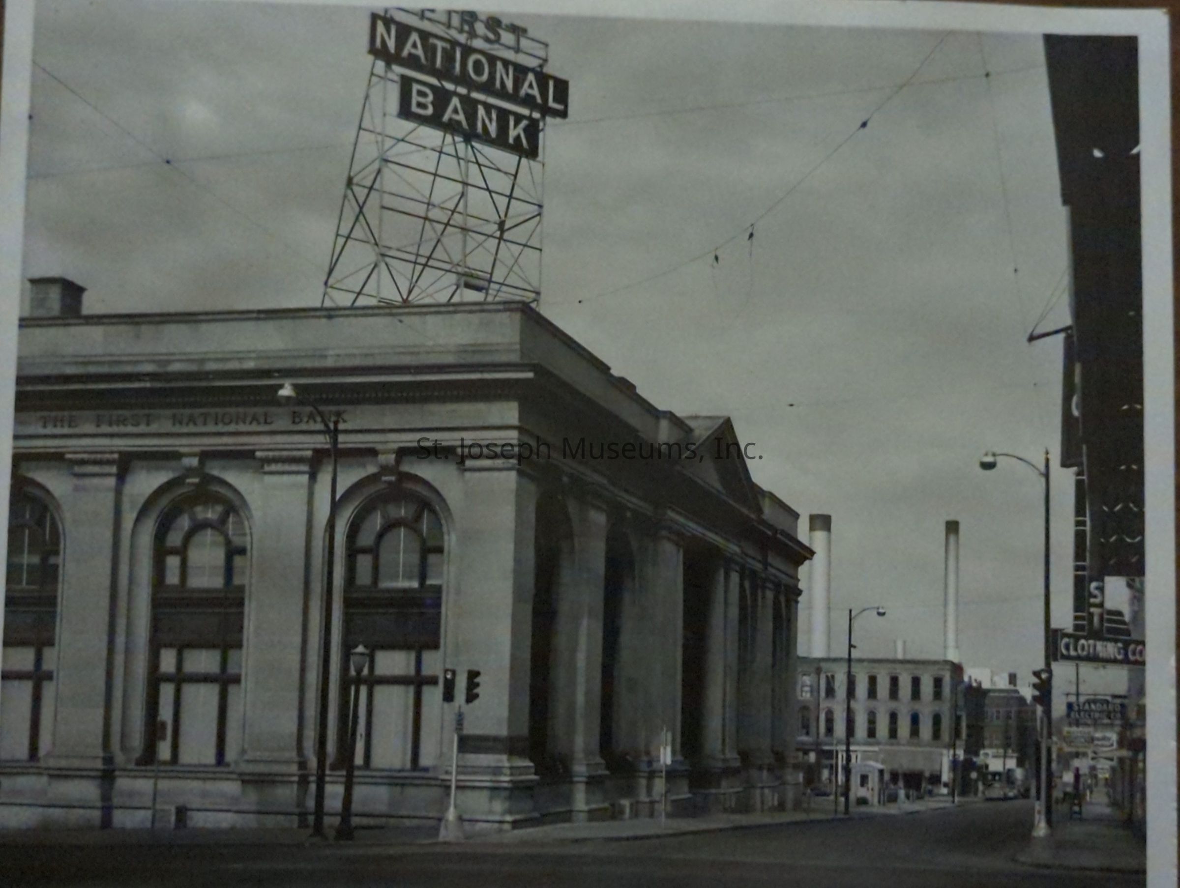 A black-and-white photo captures a historic street scene at the corner of 4th Street, looking west towards the imposing First National Bank Building. This grand structure features classical architectural details such as large, arched windows and stately columns. Above it, a prominent rooftop sign rises into the cloudy sky, emblazoned with "First National Bank" in bold letters. Streetlights line the sidewalk, and in the distance, industrial smokestacks stand tall amidst older brick buildings. The streets appear quiet.