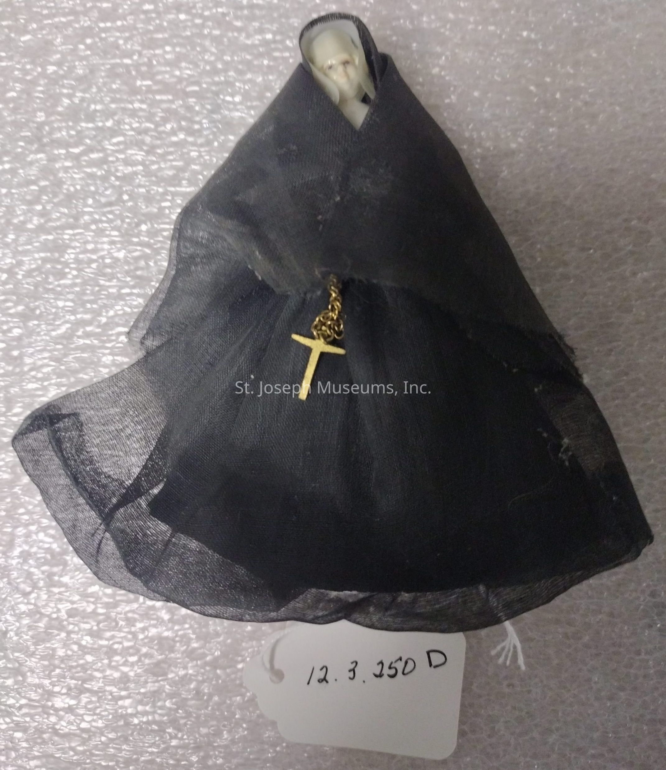 The image showcases a small figurine draped in a black fabric cloak that gives it an enigmatic and somber appearance, with a pale face peeking from beneath the hood, imparting a sense of mystery and intrigue. The adornment of a delicate golden cross hanging on a chain from the front of the cloak adds a significant detail, suggesting themes of spirituality or reverence. The background is a textured surface that enhances the focus on the figurine itself. Beneath the figure, a handwritten tag displays the numbers "12.3.250 D," which may denote a cataloging or identification system, lending an air of antique or collectible significance to the piece.