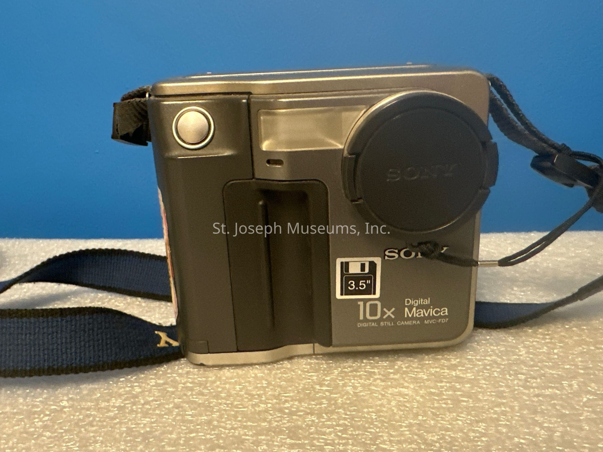 The image shows a vintage Sony Digital Mavica MVC-FD7 camera against a blue background. The camera features a prominent lens cap on the front, with the brand name "Sony" embossed on it. Additionally, there's a strap attached to the camera for ease of carrying. The device highlights its 10x optical zoom capability and uses 3.5" floppy disks for storage, indicated by the floppy disk icon on its side. The camera's finish is gray, and its design reflects a sturdy build typical of 1990s models, capturing the essence of early digital photography technology.