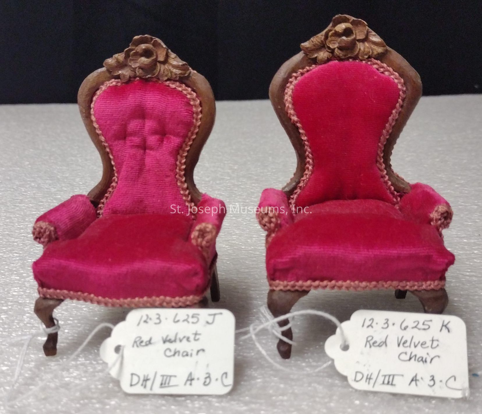 The image features two intricately designed miniature chairs, both upholstered in rich red velvet fabric. Each chair has a high, rounded back adorned with ornate wood carvings resembling floral motifs at the top. The plush velvet not only covers the seat and back but also extends to the armrests, creating a luxurious appearance. The chairs are set against a neutral backdrop, highlighting their detailed craftsmanship. Attached to each chair is a small white tag with handwritten notes, identifying them as "Red Velvet Chair," along with some code numbers likely related to cataloging or inventory purposes. These tags are fastened with thin string loops around the legs of the chairs. The vivid red color contrasts with the dark wood frame, emphasizing the classic and elegant design typical of vintage furniture miniatures. The chairs symbolize refinement and attention to detail, making them visually captivating as collectible pieces or elements of a detailed dollhouse setting.