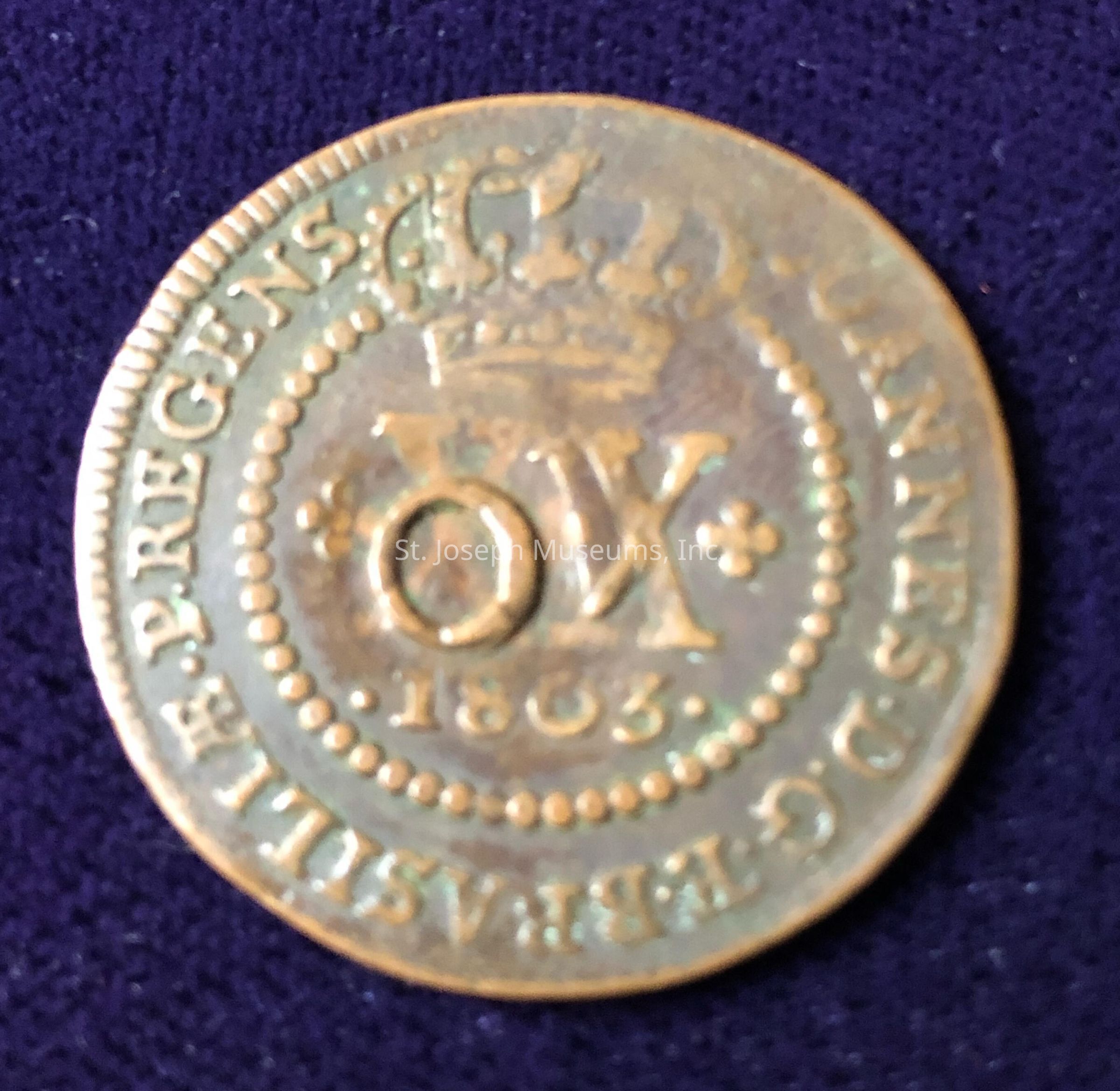 The image displays a coin featuring intricate details, including a prominent crown at the top and the year "1803" engraved below. In the center, there is a large "OX" surrounded by decorative elements and flanked by what appears to be stars. The outer edge of the coin is inscribed with a series of words, potentially in Latin, which are partially visible and include phrases such as "E.P. REGENS" and "DUCANNES." The coin, with its aged patina and metallic sheen, rests on a dark surface, perhaps made of fabric, which contrasts with its weathered bronze appearance, highlighting the craftsmanship and historical significance evident in its design.