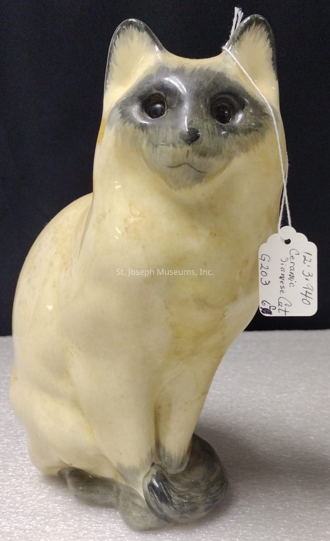 The image depicts a ceramic statue of a cat that resembles a Siamese breed, characterized by its light-colored body and darker accents on the face, ears, and tail. The figure is crafted with smooth surfaces and subtle shading to emphasize the contours and expression of the feline, giving it a somewhat realistic yet stylized appearance. The cat is sitting upright with its tail wrapped around its paws, and a small tag is attached, featuring handwritten information about the ceramic piece. The background is a plain dark surface, allowing the details and form of the ceramic cat to stand out prominently in the image.