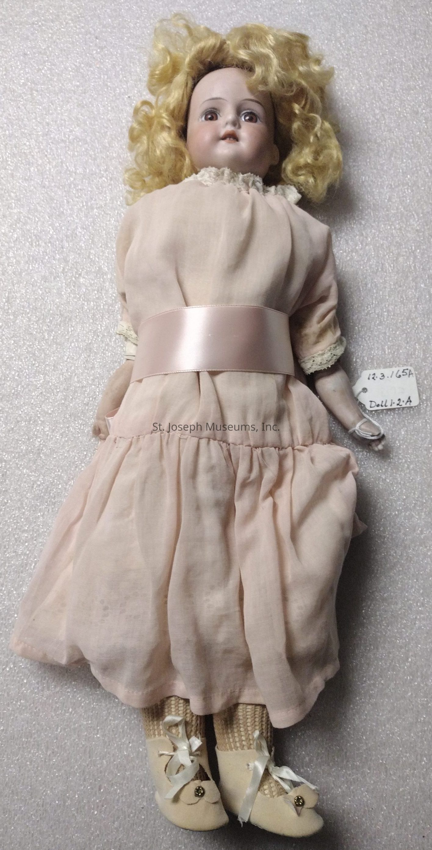 The image depicts a vintage porcelain doll with curly blonde hair, wearing a delicate pale pink dress adorned with lace trimming and a satin sash around the waist. The doll's face features painted details that include rosy cheeks and lips, as well as blue eyes gazing upwards, contributing to its classic, antique look. It is dressed elegantly, combining old-fashioned charm with intricate craftsmanship, evident in the refined texture of its clothing and the svelte shape of its limbs. Completing the attire, the doll wears knitted socks and soft-looking cream-colored shoes decorated with ribbon ties and small button accents. A small tag attached to its wrist displays the numbers "12.3.165" and the label "Doll 1.2:A," suggesting it might be part of a curated collection or inventory. The entire presentation is set against a softly-textured, neutral background, which focuses attention on the exquisite details of the doll, allowing its nostalgic and historical essence to stand out vividly in the frame.