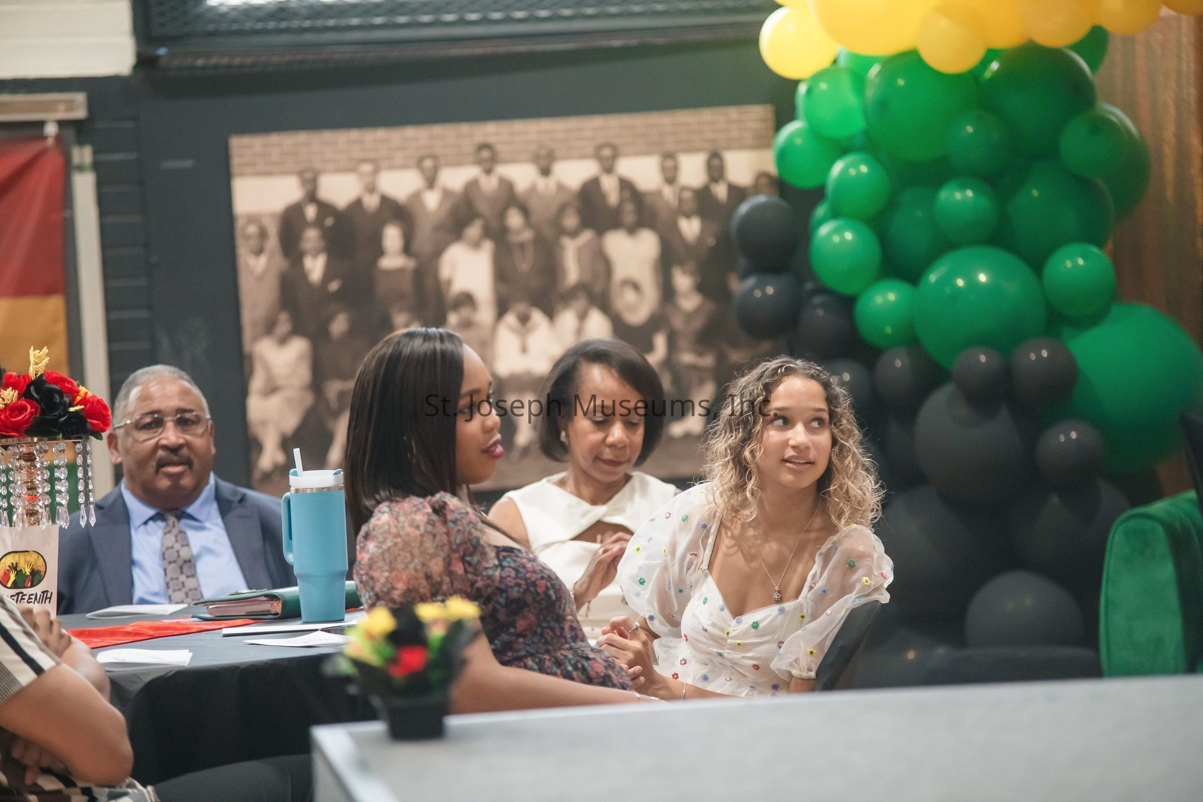 The image depicts several people seated around a table at what appears to be a formal or celebratory event, characterized by decorations of colorful balloons in green, yellow, and black. In the foreground, there are flowers and a decorative centerpiece with red accents, creating a vibrant atmosphere. Among the individuals, one man is seen wearing a suit and tie, while two women are dressed in elegant attire, suggesting that they are attending an important gathering. The backdrop includes a large, sepia-toned group photograph, adding a historical or commemorative element to the setting. The attendees seem engaged and attentive, contributing to a sense of community and togetherness.